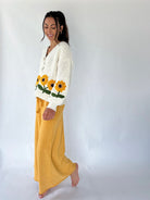 A woman modeling a cream knit cardigan with yellow and green sunflower embroidery on the front and sleeves, paired with mustard yellow pants, against a plain white studio background.