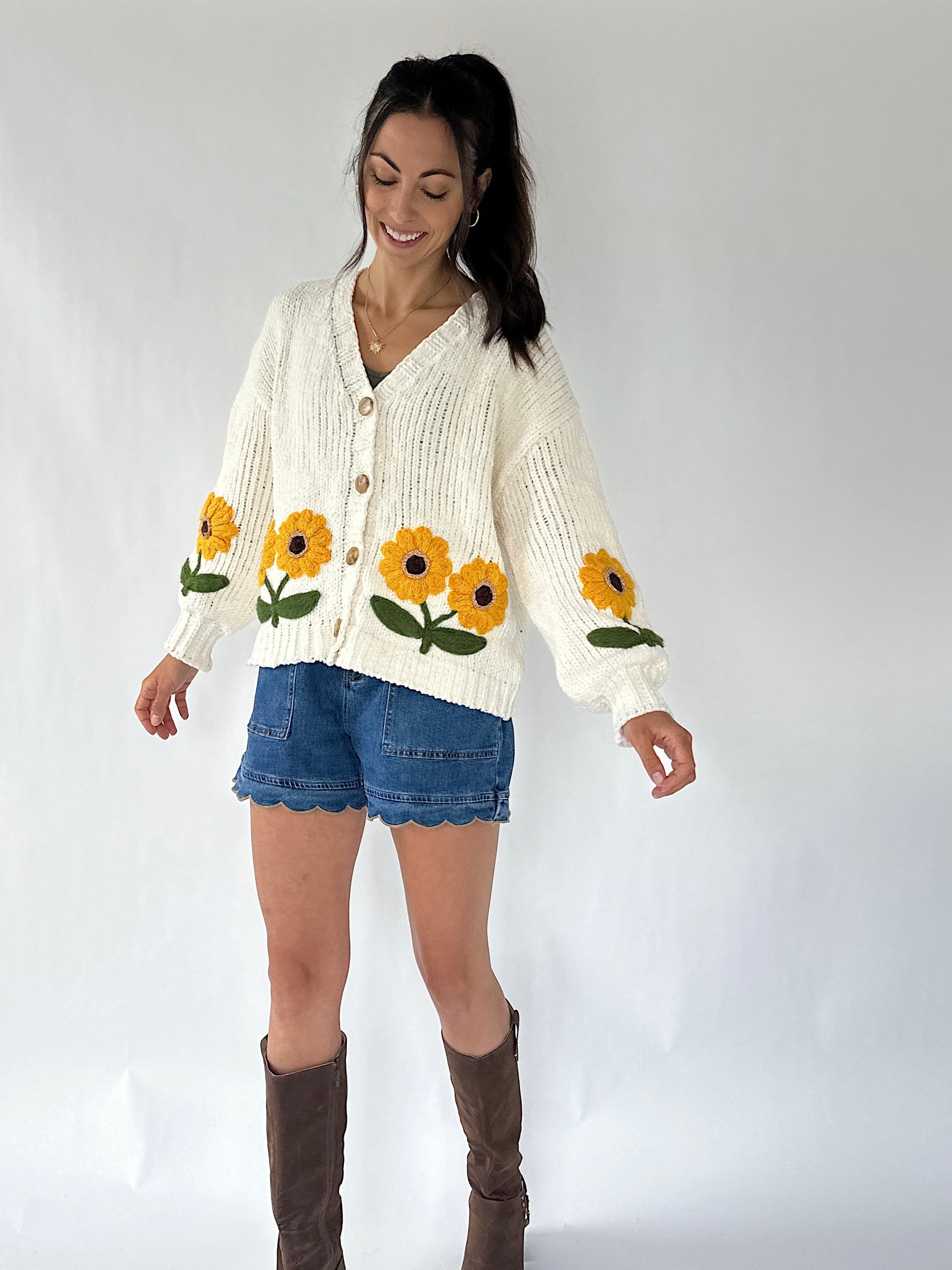 A woman modeling a cream-colored cable-knit cardigan sweater with yellow sunflower and green leaf embroidery on the sleeves and hem, paired with denim shorts and brown boots against a white studio background.