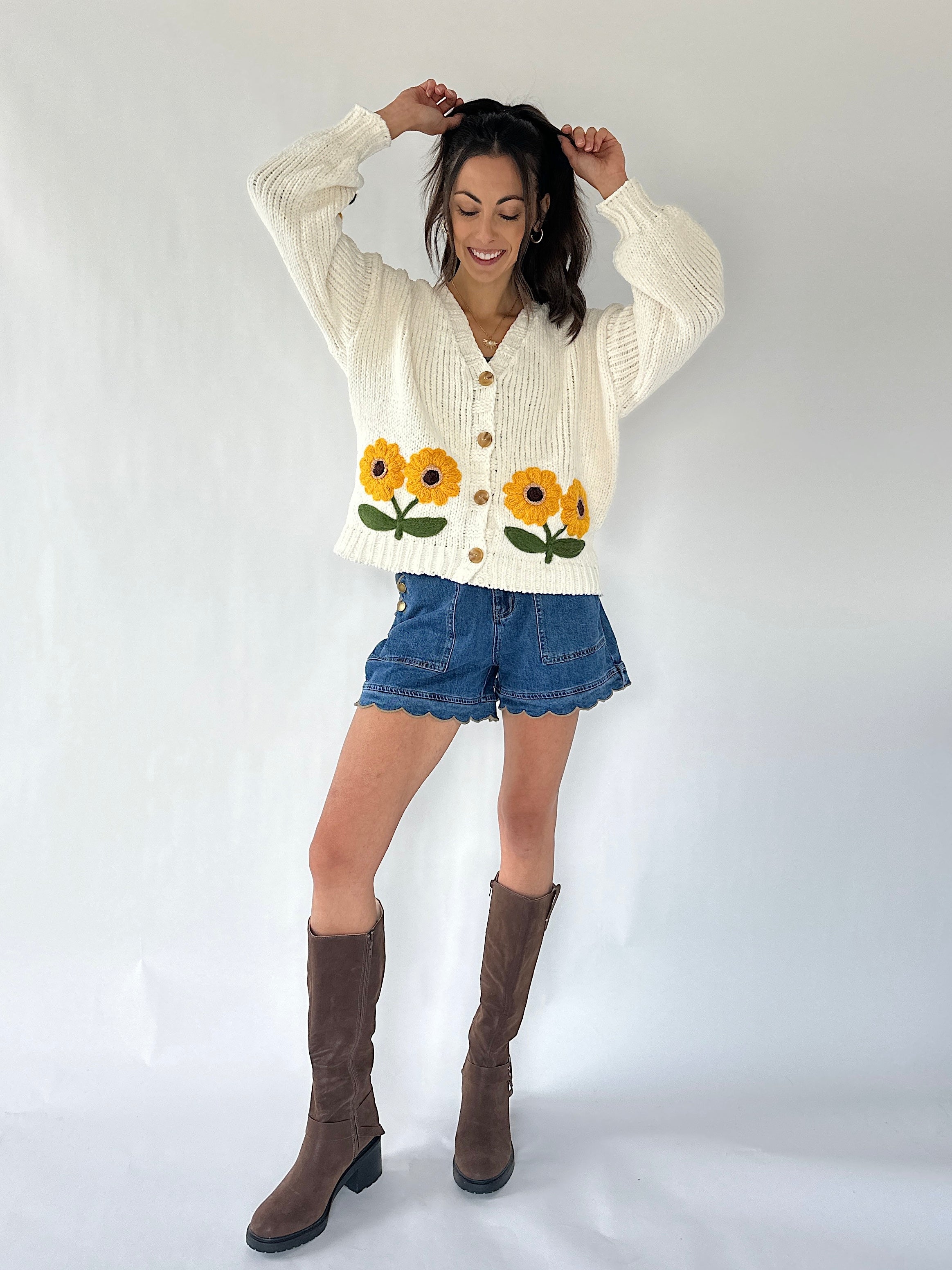A woman modeling a cream-colored cable-knit cardigan sweater with embroidered sunflowers, paired with denim shorts and brown knee-high boots, against a plain white studio background.