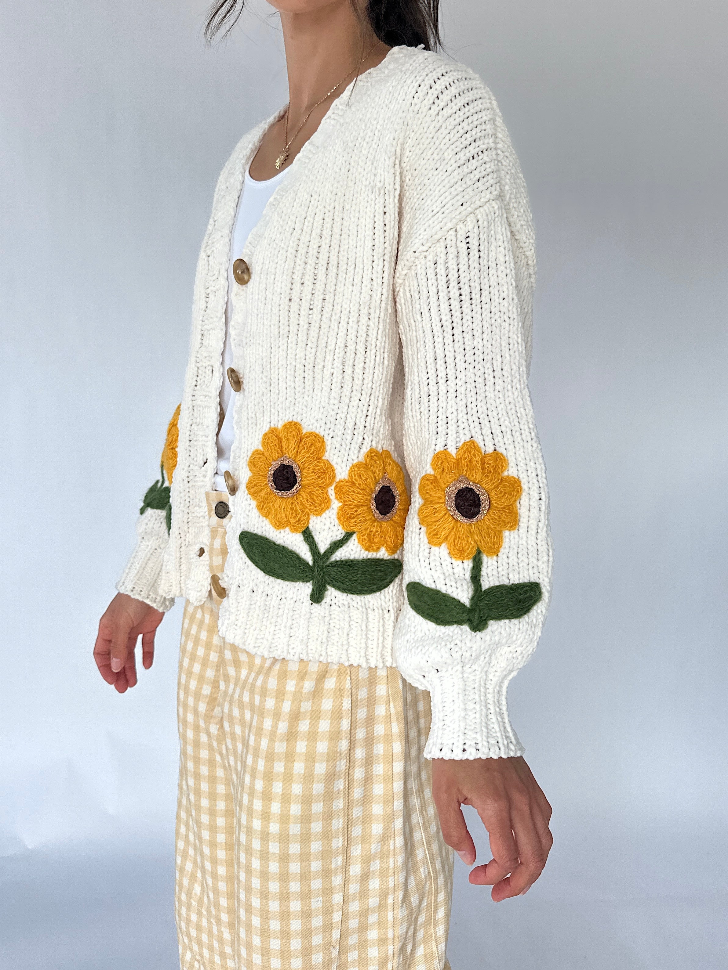A woman wearing a cream-colored chunky knit cardigan with large yellow sunflower patches on the front and sleeves, paired with a white top and yellow-and-white gingham pants, set against a plain light gray background.