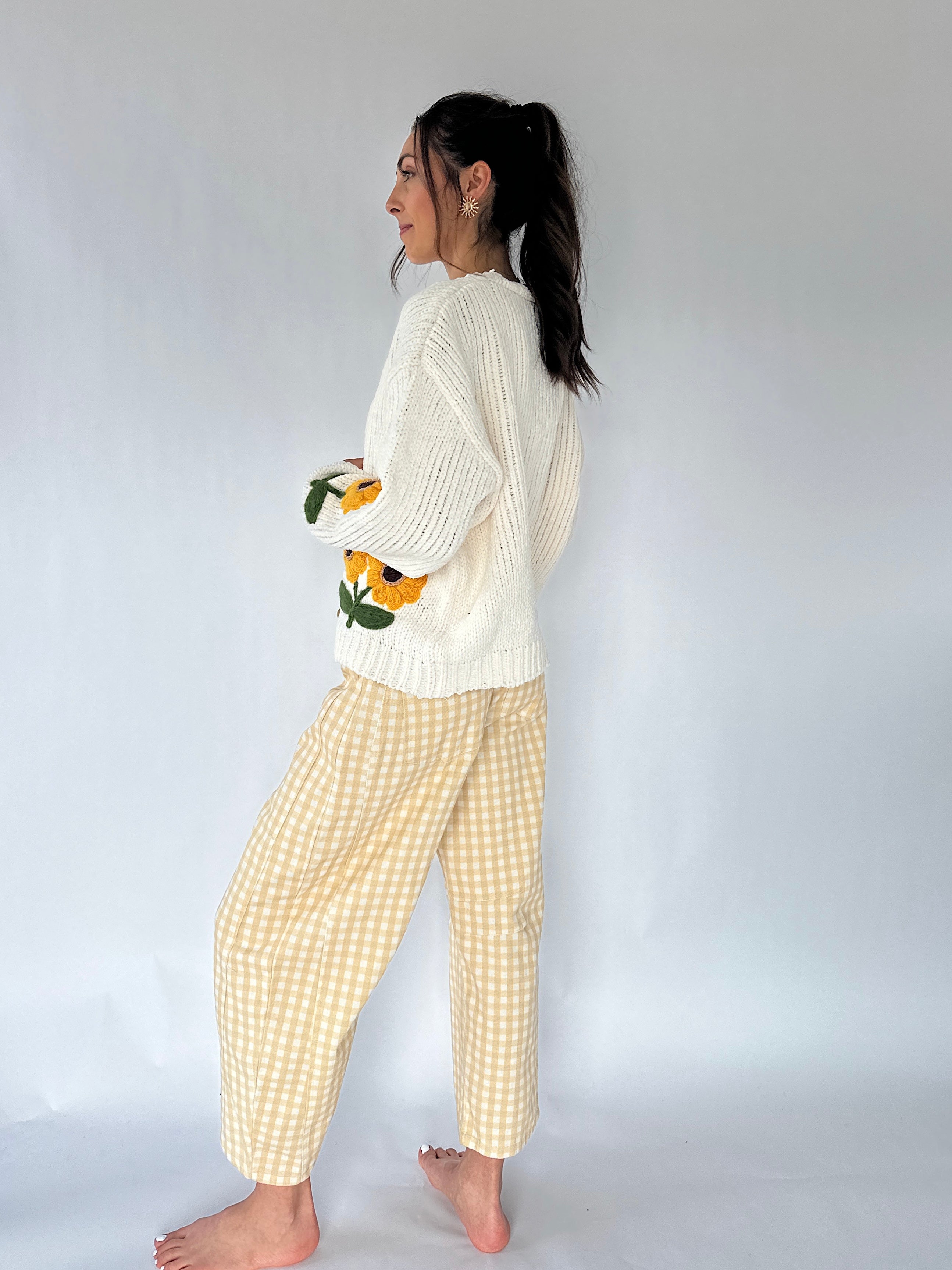 A woman stands barefoot in profile wearing a cream ribbed knit sweater with yellow sunflower embroidery on the sleeves and light beige-and-white gingham cropped pants, against a plain light gray studio background.