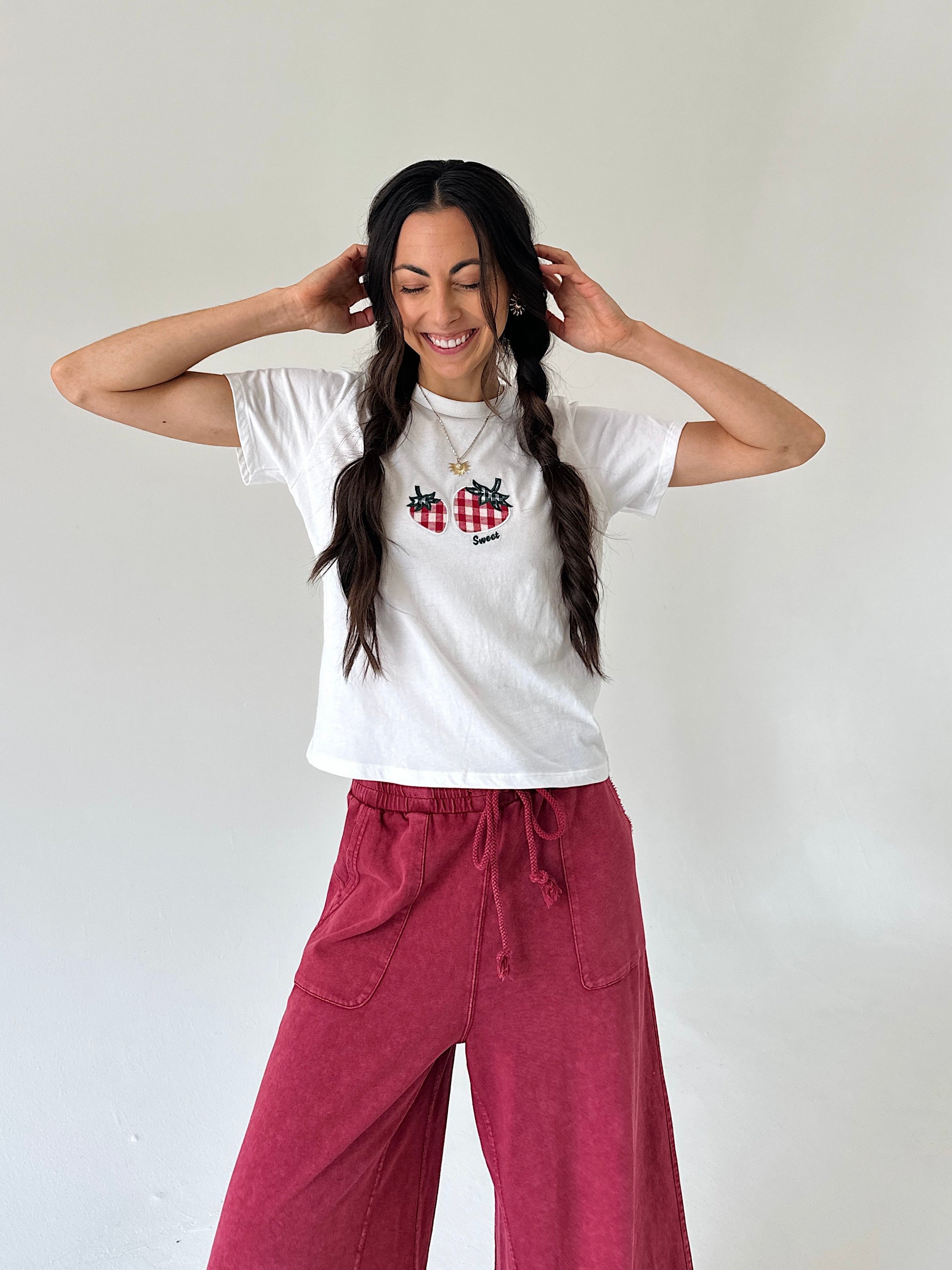 A woman with long dark hair in two braids is smiling and posing in a white crewneck t-shirt with a red-and-black plaid strawberry graphic, paired with dusty rose wide-leg pants, against a plain off-white wall.