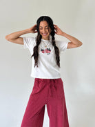 A woman with long dark hair in two braids is smiling and posing in a white crewneck t-shirt with a red-and-black plaid strawberry graphic, paired with dusty rose wide-leg pants, against a plain off-white wall.