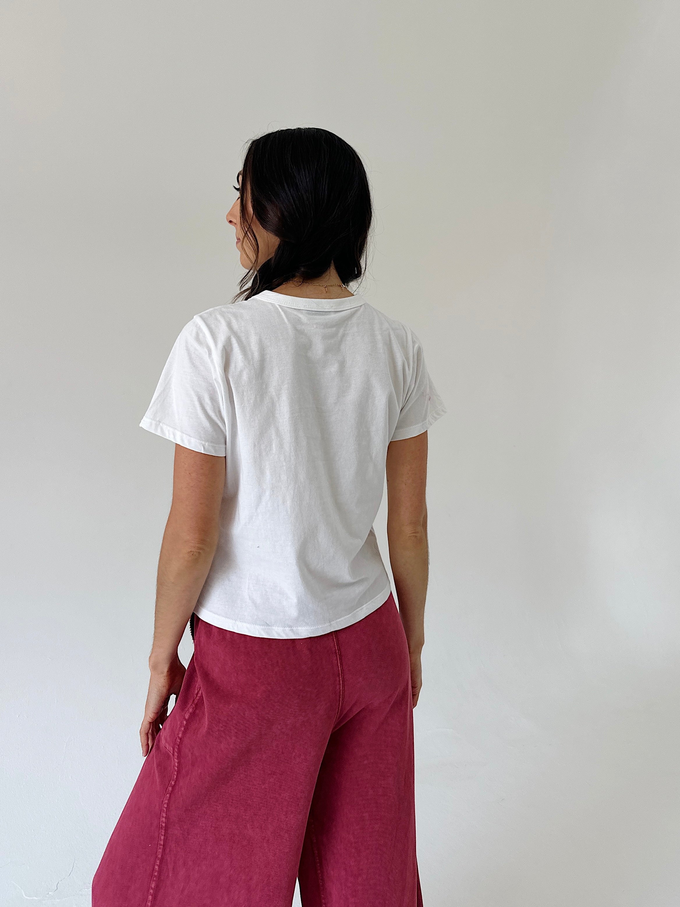 A woman shown from behind wearing a white short-sleeve t-shirt and dusty rose wide-leg pants, standing against a plain off-white wall.
