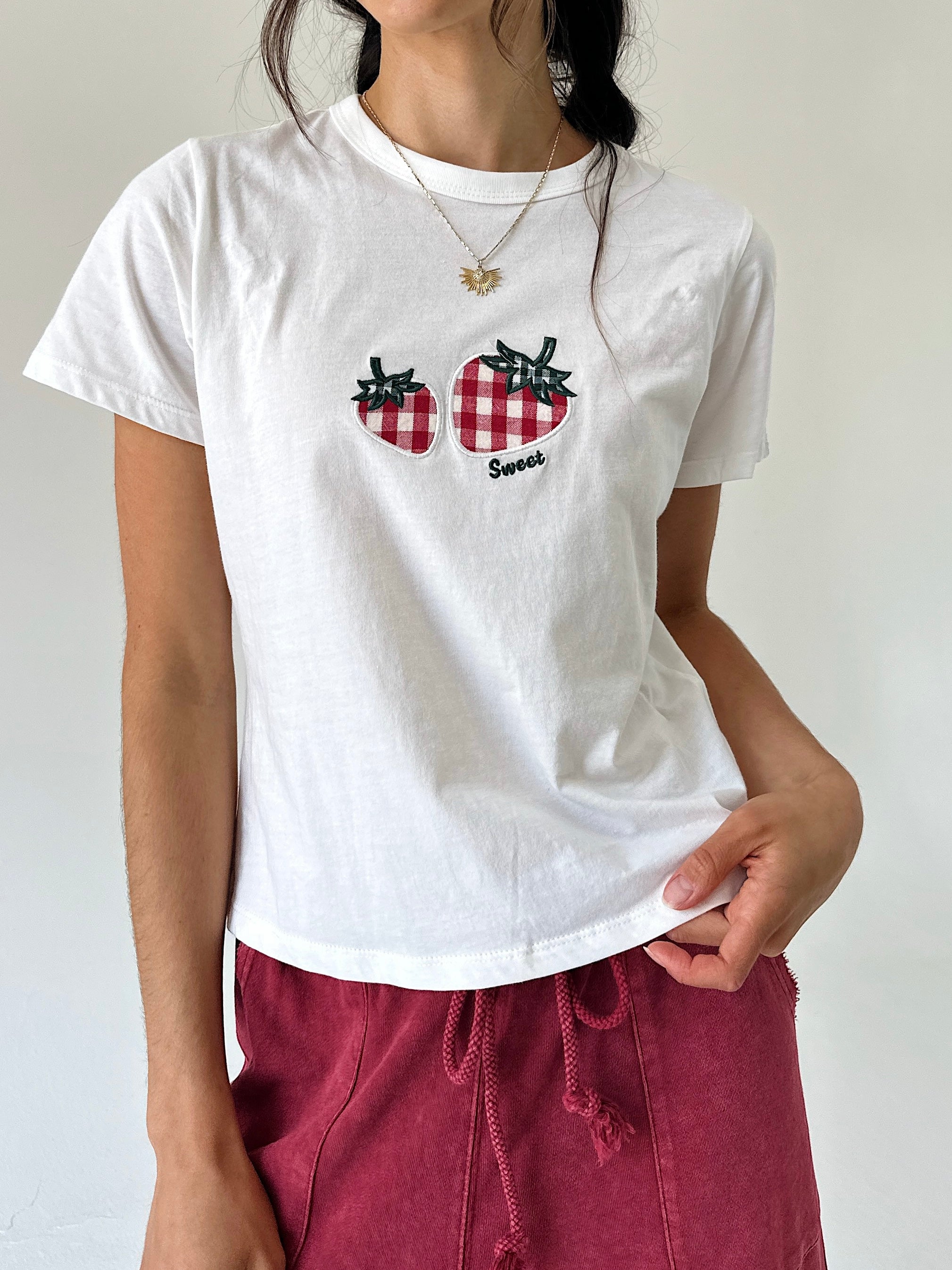 A woman wearing a white short-sleeve crewneck t-shirt with a red and black gingham embroidered strawberry graphic on the chest, paired with burgundy drawstring pants.
