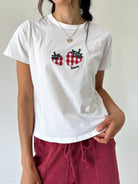 A woman wearing a white short-sleeve crewneck t-shirt with a red and black gingham embroidered strawberry graphic on the chest, paired with burgundy drawstring pants.