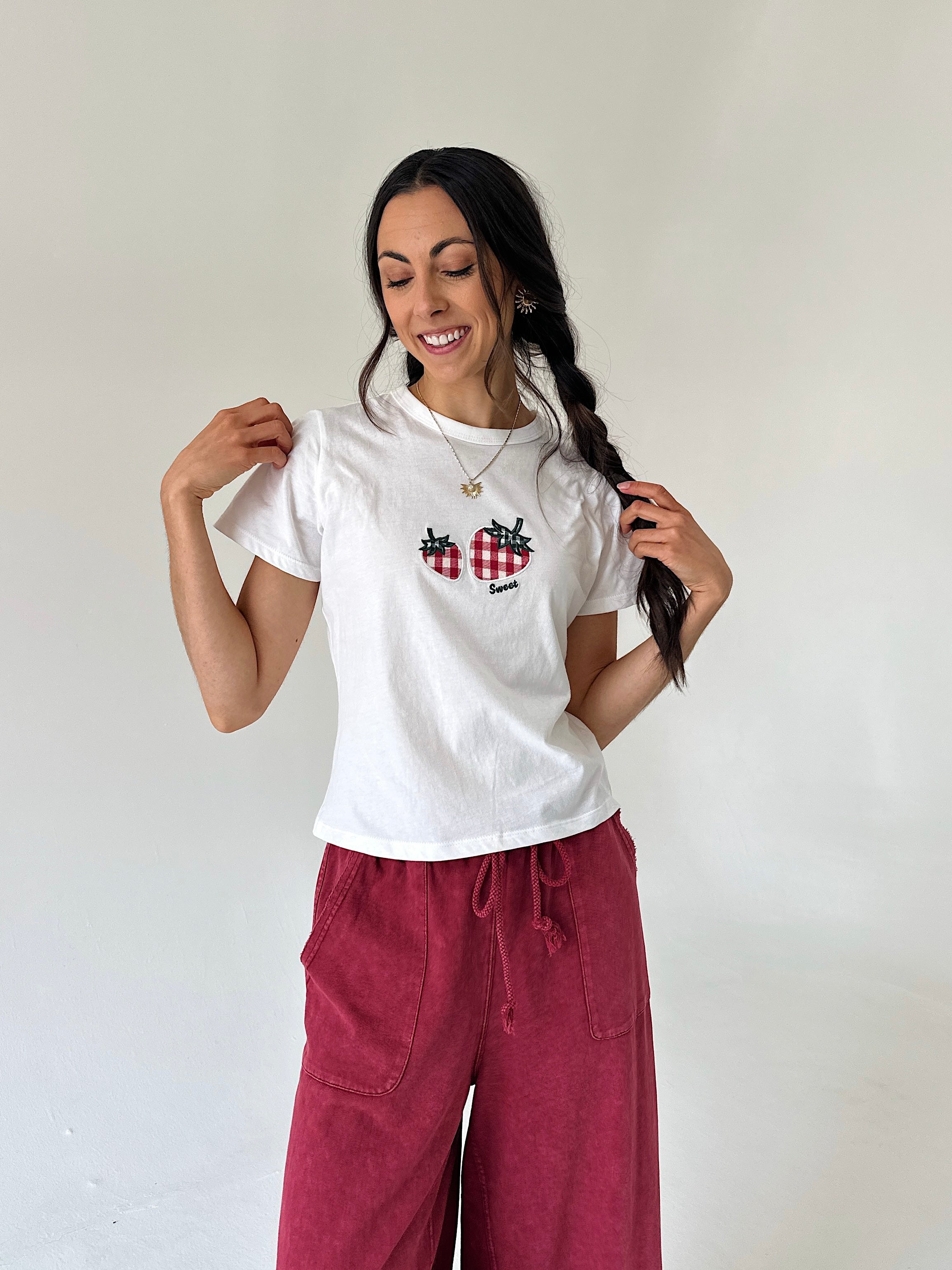 A woman modeling a white short-sleeve t-shirt with a red-and-white checkered strawberry graphic and a small black "sweet" lettering, paired with burgundy wide-leg pants, against a plain off-white wall.