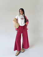 A woman modeling a white graphic t-shirt, wide-leg burgundy wide leg sweatpants, a striped knit cardigan, and beige suede shoes against a plain light gray studio background.