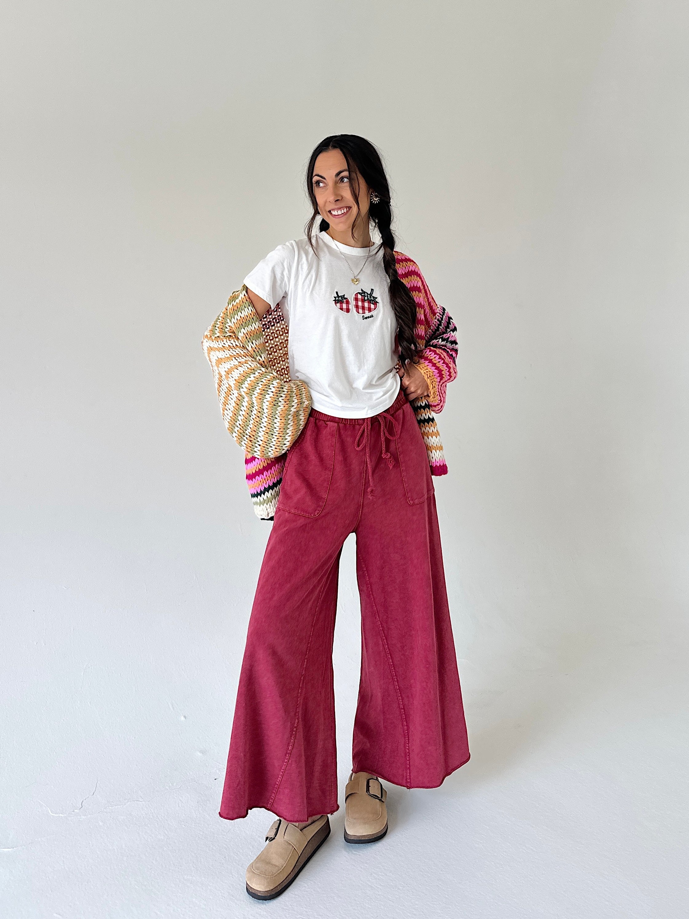 A woman modeling a white graphic t-shirt, wide-leg burgundy wide leg sweatpants, a striped knit cardigan, and beige suede shoes against a plain light gray studio background.