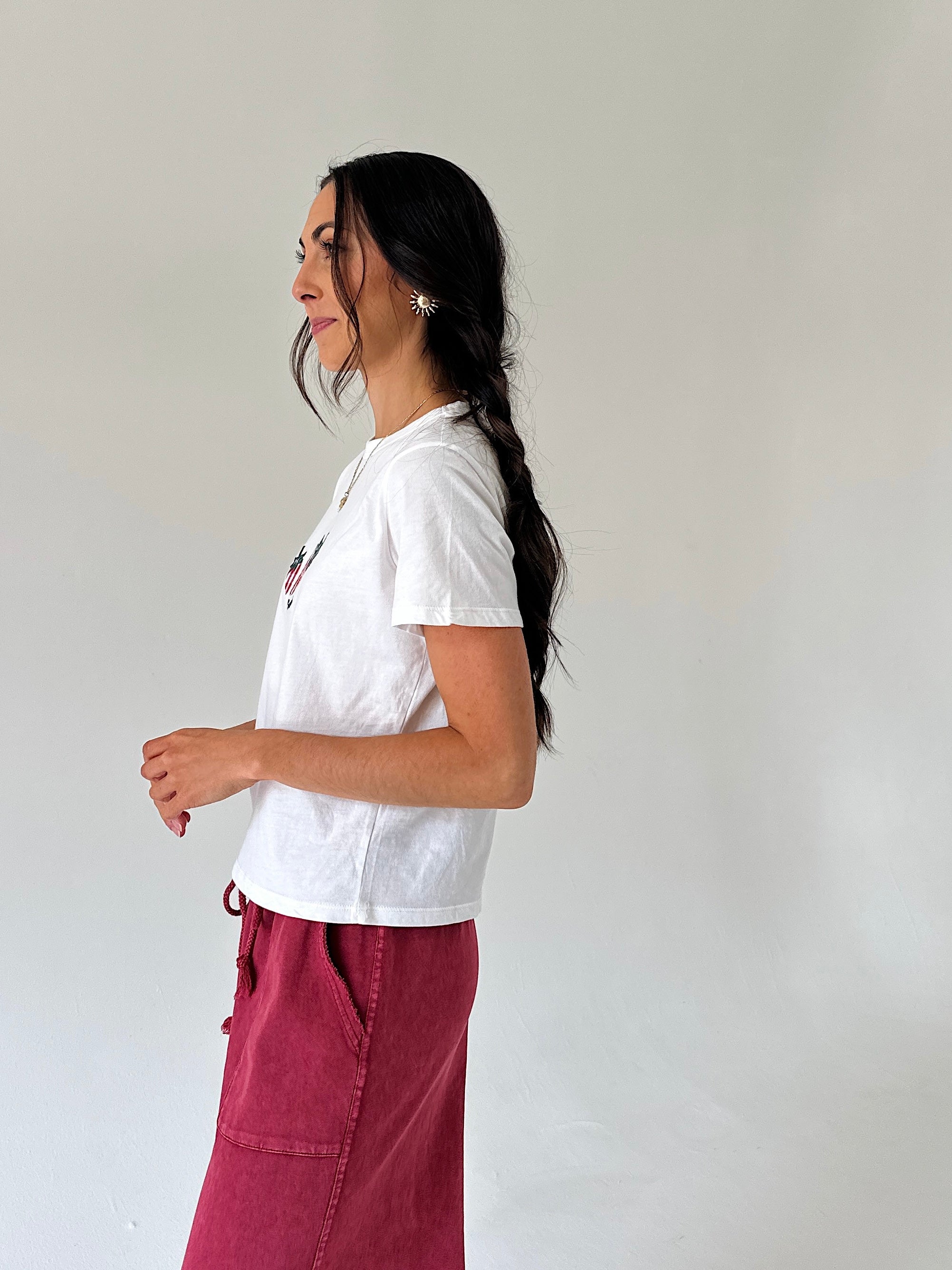 A woman with long dark hair in a side-profile pose, wearing a white short-sleeve t-shirt and a dusty boho red pants, standing against a plain off-white wall.