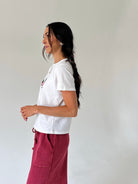 A woman with long dark hair in a side-profile pose, wearing a white short-sleeve t-shirt and a dusty boho red pants, standing against a plain off-white wall.