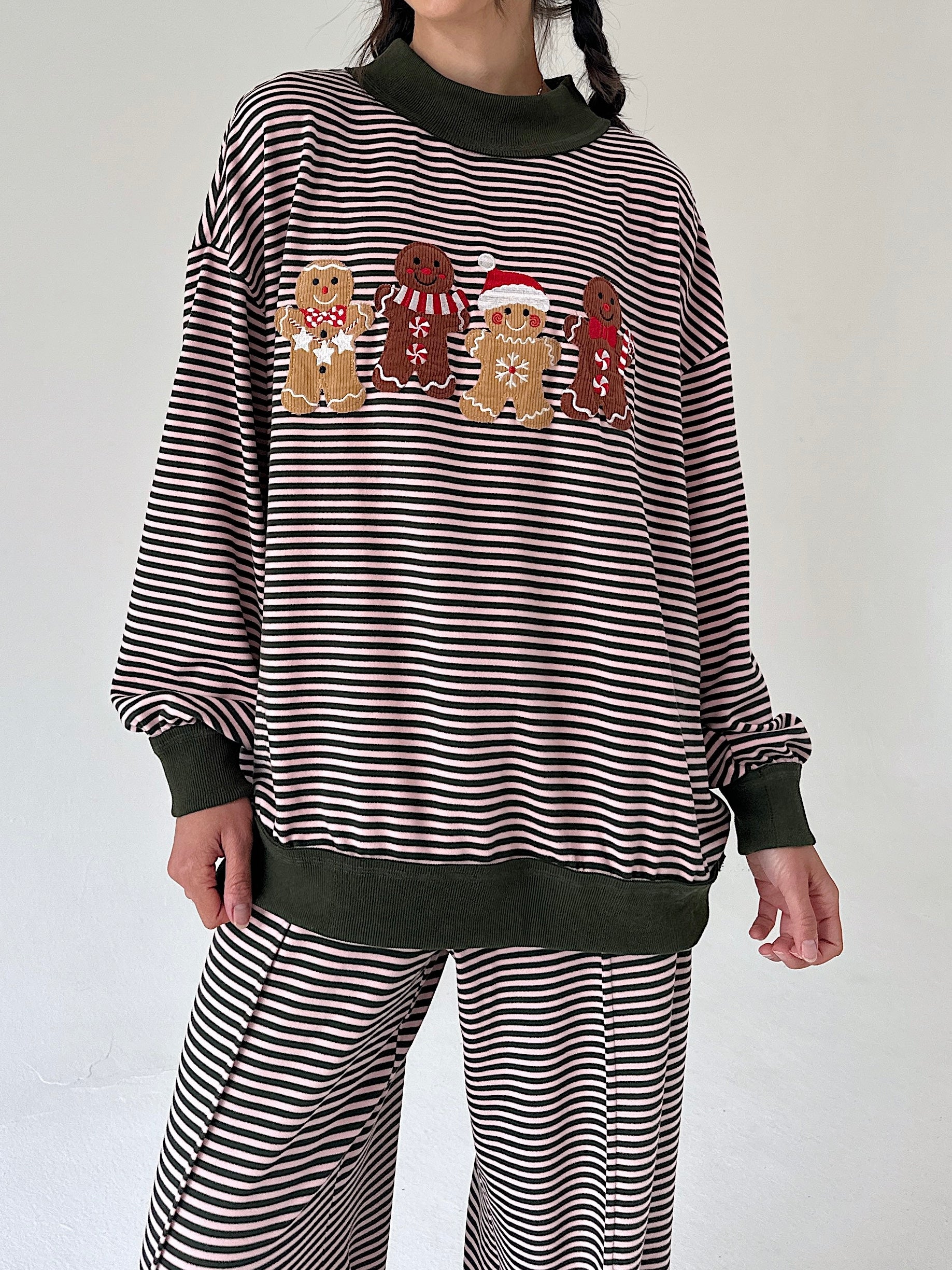 A person wearing a pink and dark green horizontally striped long-sleeve sweatshirt with four gingerbread cookies across the chest, paired with matching striped pants. The sweatshirt has dark green ribbed trim at the collar, cuffs, and hem, and the gingerbread cookies feature red and green accents.