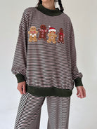 A person wearing a pink and dark green horizontally striped long-sleeve sweatshirt with four gingerbread cookies across the chest, paired with matching striped pants. The sweatshirt has dark green ribbed trim at the collar, cuffs, and hem, and the gingerbread cookies feature red and green accents.