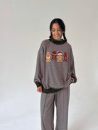 A woman modeling a matching holiday pajama set consisting of a long-sleeve top and pants with horizontal stripes and gingerbread cookie graphics on the chest, set against a plain white studio background.