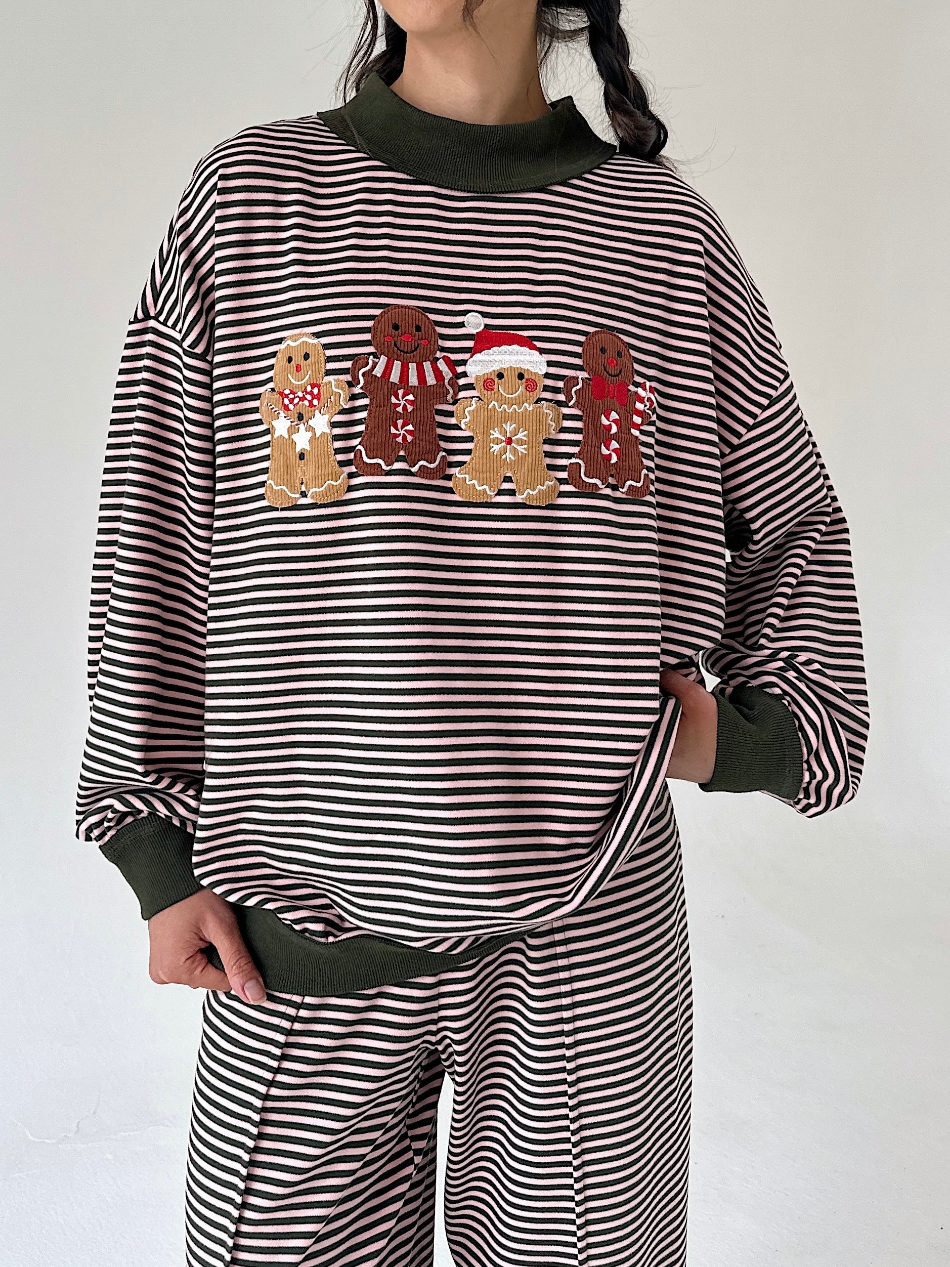 A person wearing a horizontally striped long-sleeve sweatshirt with a festive gingerbread man graphic across the chest, paired with matching striped pants. The garment features solid dark olive green ribbed mock neck, cuffs, and hem.