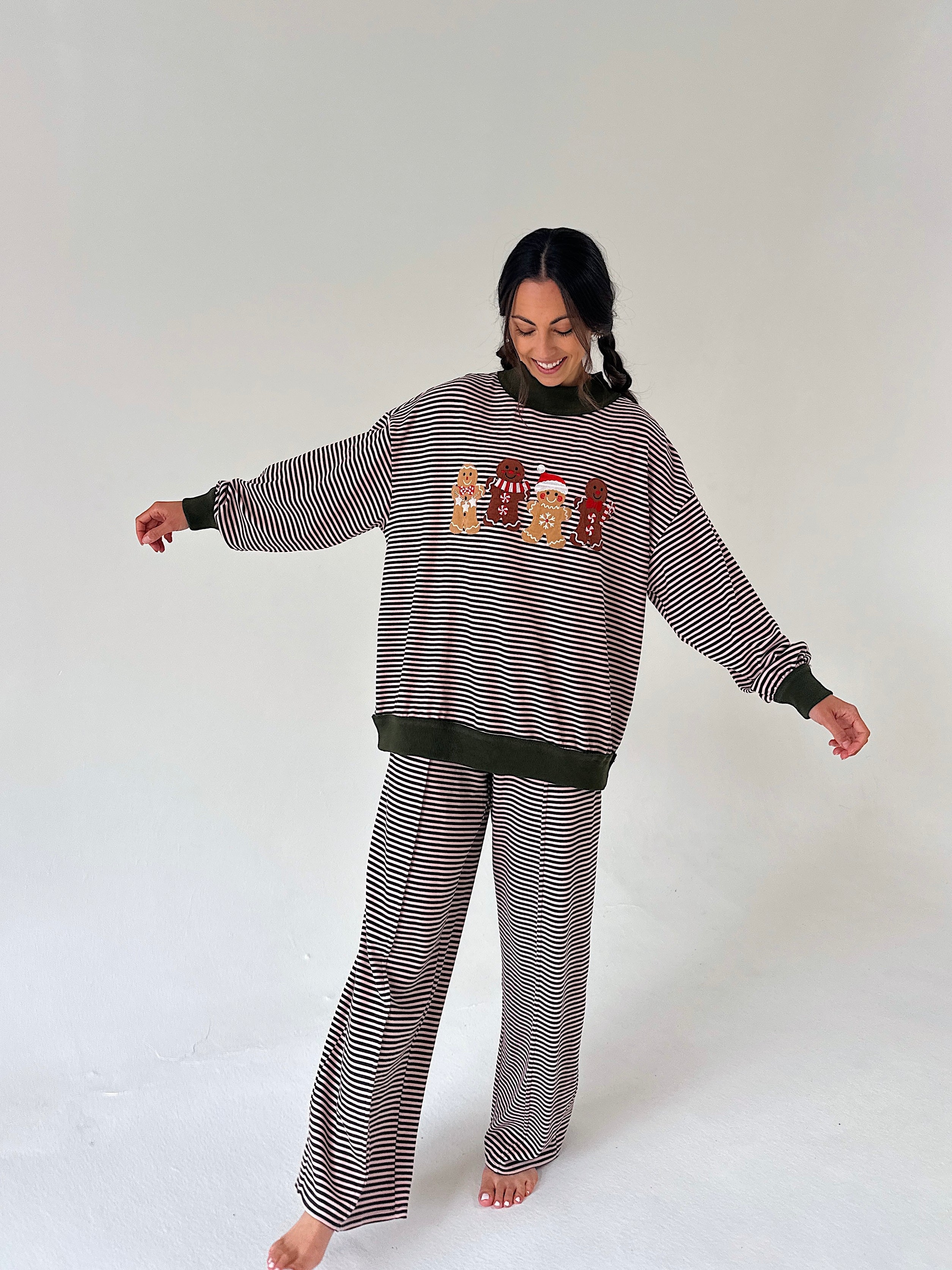 A woman modeling a matching holiday-themed loungewear set consisting of a long-sleeve striped sweatshirt and wide-leg pants. The set features thin pink and dark green stripes with a dark olive green ribbed hem, cuffs, and collar.