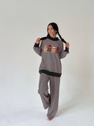 A woman modeling a matching two-piece loungewear set consisting of a long-sleeve striped sweater and wide-leg pants. The set features thin horizontal stripes, solid black ribbed trim at the collar, cuffs, and hem, and a central graphic of three cartoon characters wearing red hats.