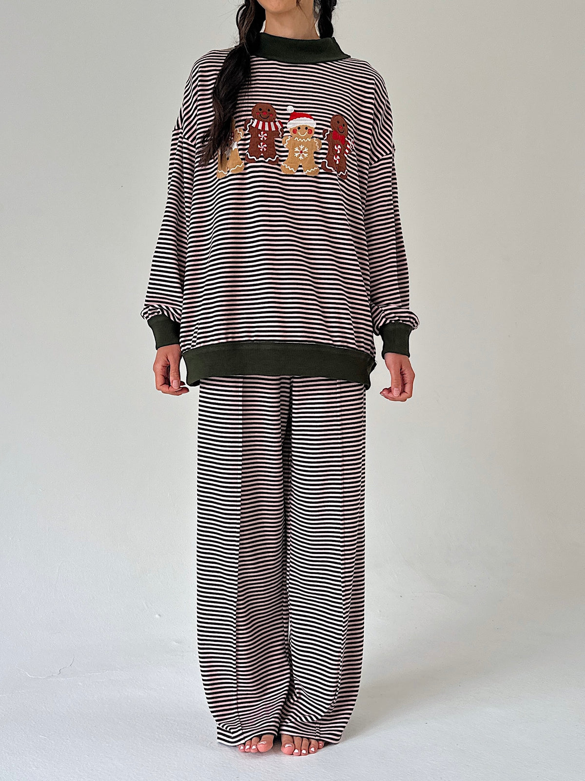A model wearing a matching two-piece loungewear set consisting of a long-sleeve top and wide-leg pants, both featuring horizontal stripe pattern. The top has a black crew neck, ribbed cuffs and hem, and a chest graphic of three gingerbread cookies wearing Santa hats.