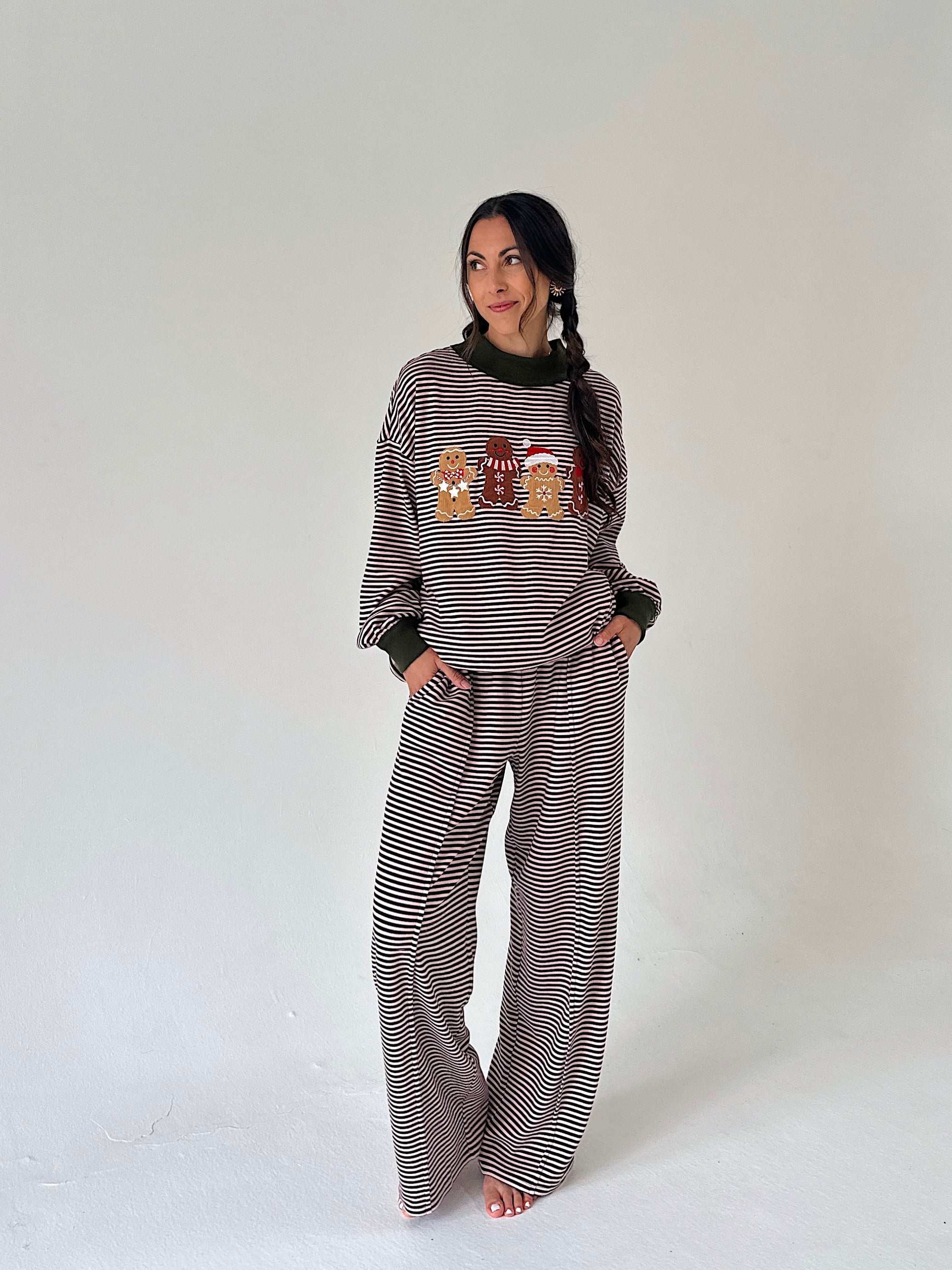A woman modeling a matching holiday-themed loungewear set consisting of a long-sleeve top with a gingerbread cookie graphic and striped pattern, and matching wide-leg pants, against a plain light gray background.