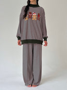 A model wearing a matching two-piece loungewear set consisting of a long-sleeve top and wide-leg pants, both featuring a horizontal stripe pattern. The top has a black crew neck, ribbed cuffs and hem, and a chest graphic of three gingerbread cookies wearing Santa hats.
