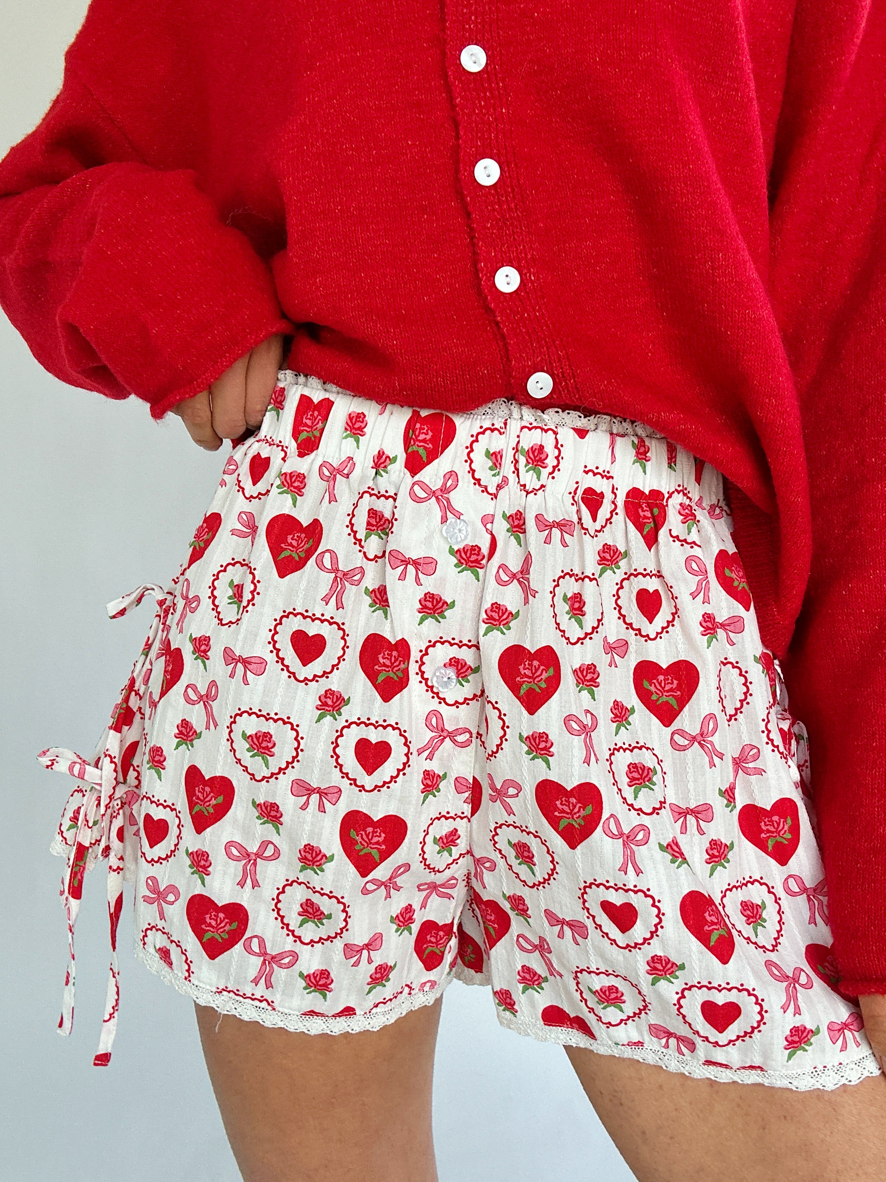 Person wearing a red cardigan and white shorts with red heart and bow pattern on a light gray background