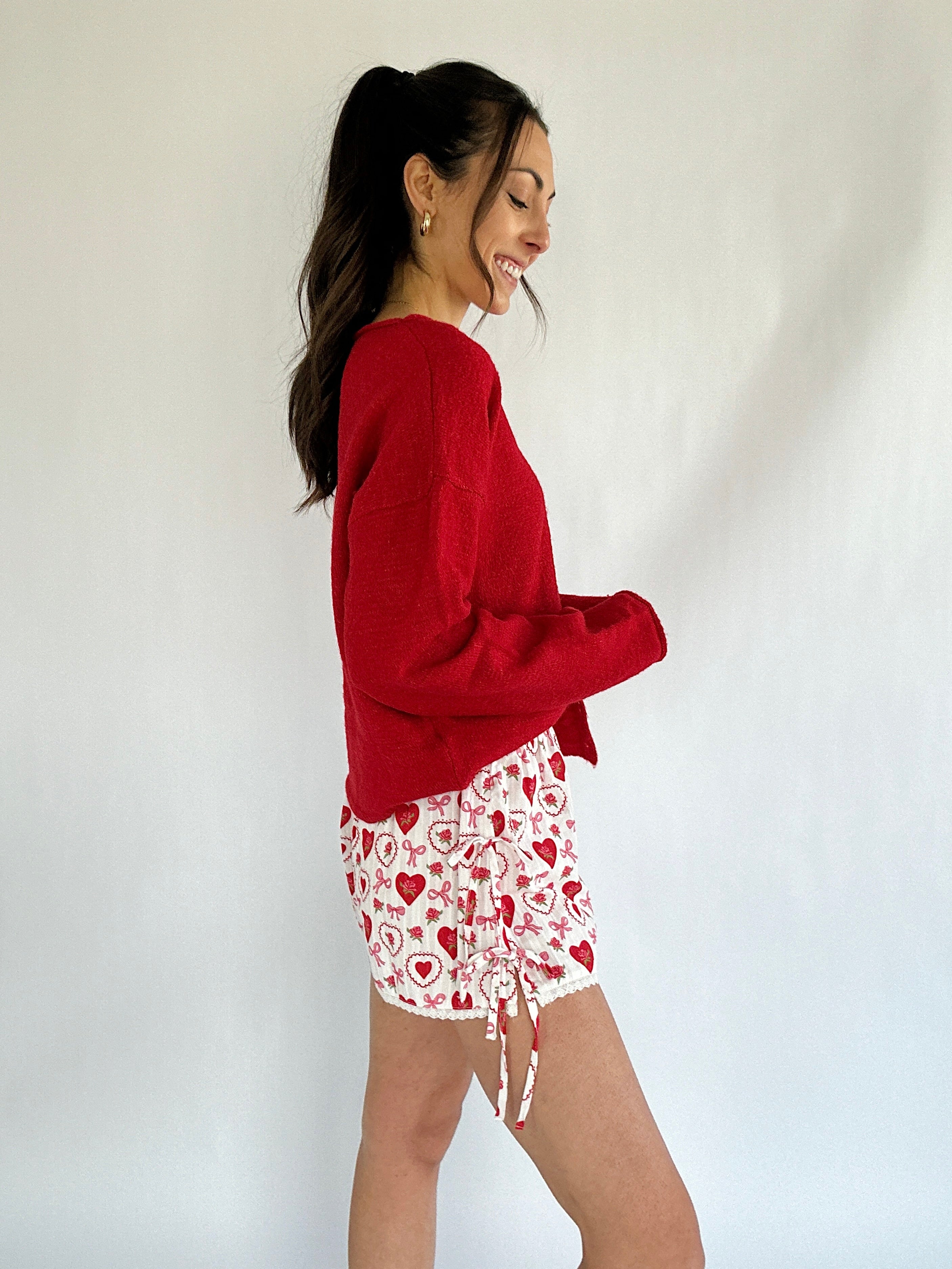 Woman wearing a red sweater and white shorts with red heart patterns on a plain background from side