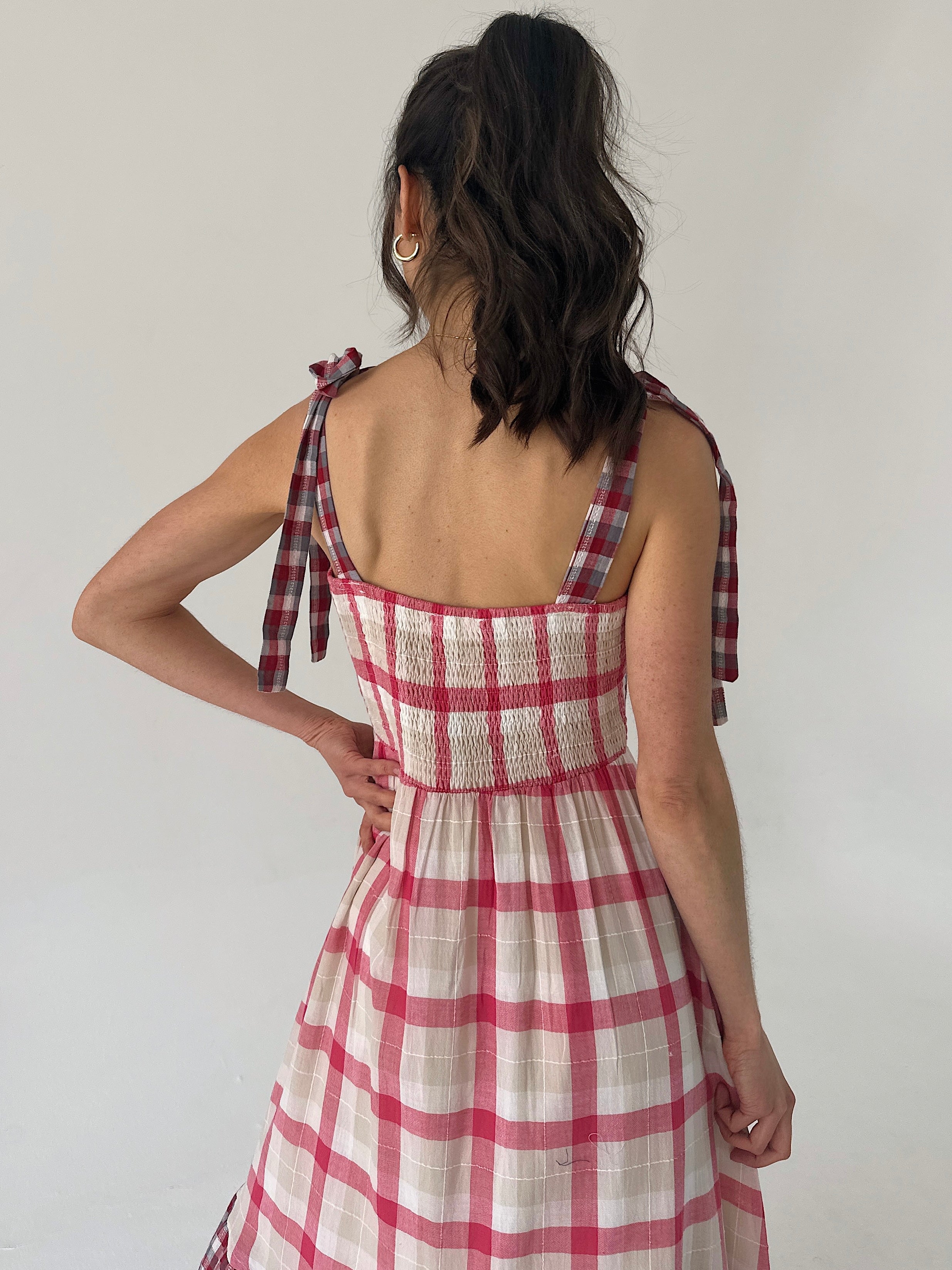 Woman wearing a red and white checkered dress with bow straps and smocked back on a plain background