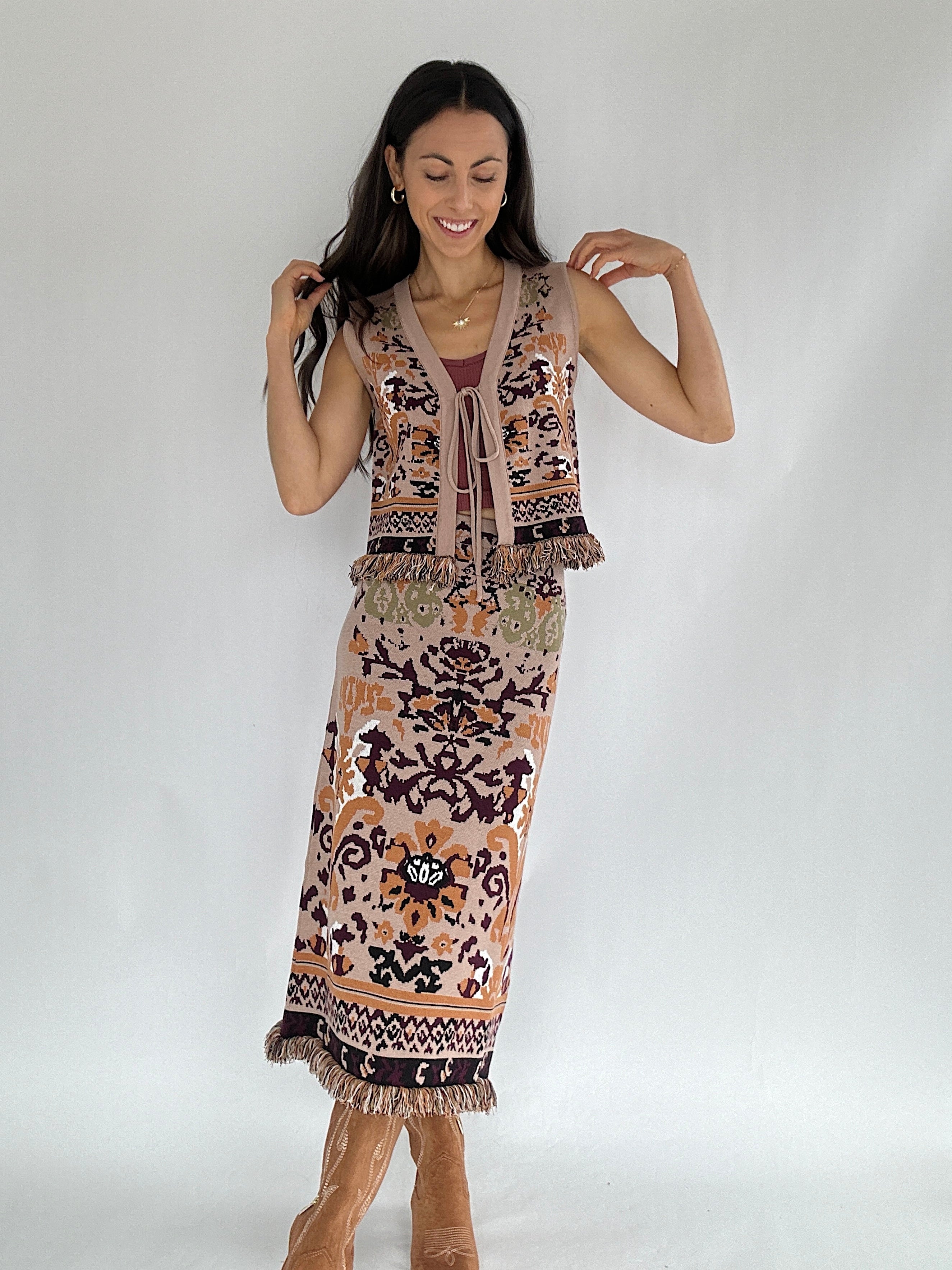 Woman wearing a boho patterned knit vest with tie front and matching knit skirt with a plain background
