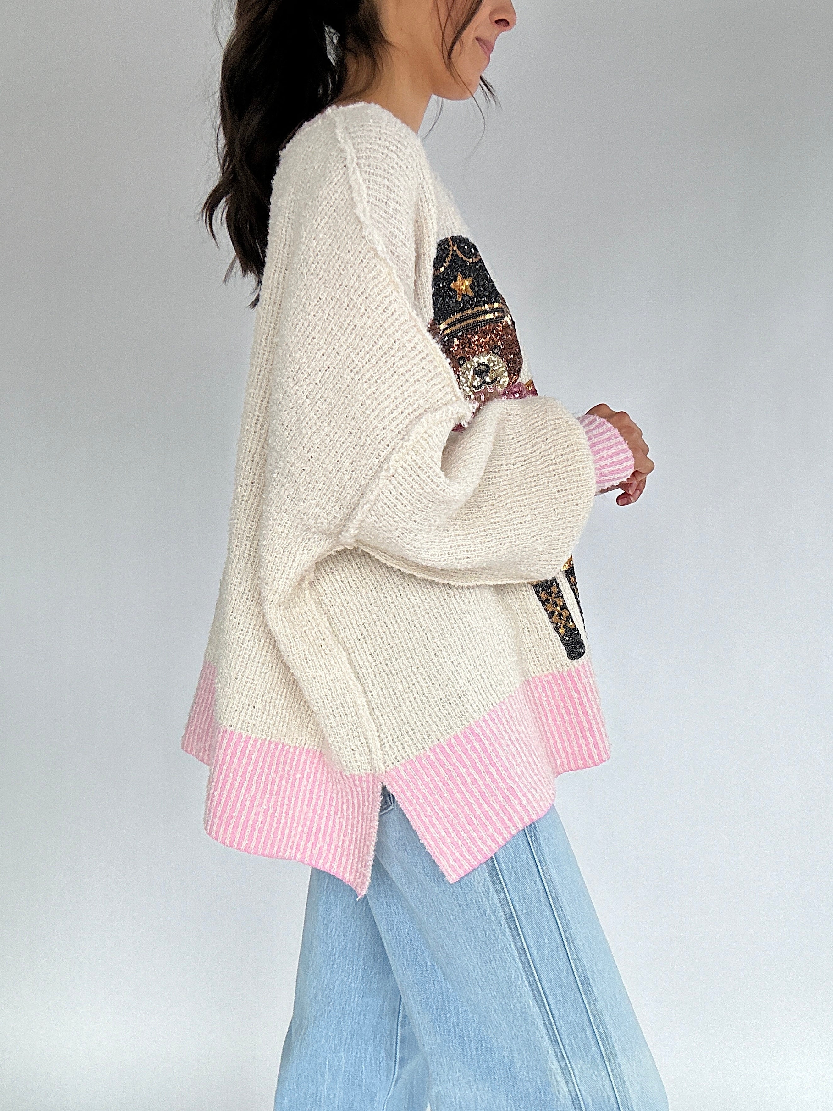 Person wearing a cream teddy sequin sweater with pink trim and jeans on a plain background to show side view