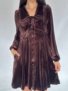 Brown velvet mini dress with peter pan collar, tie detail, button front and side pockets worn by a person on a white background