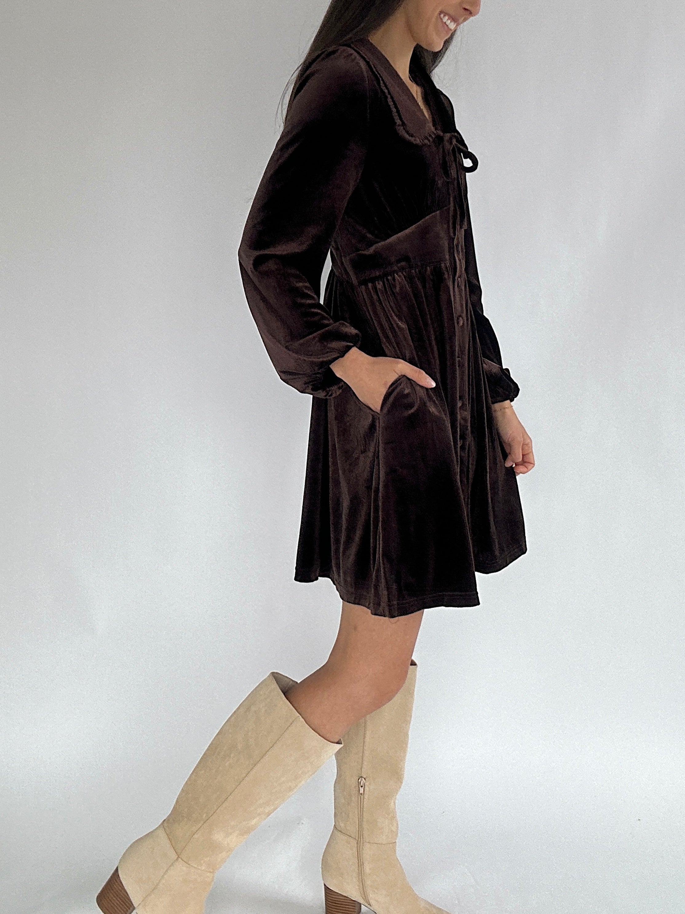 Person wearing a brown velvet mini dress with beige knee-high boots on a white background with hands in side pockets
