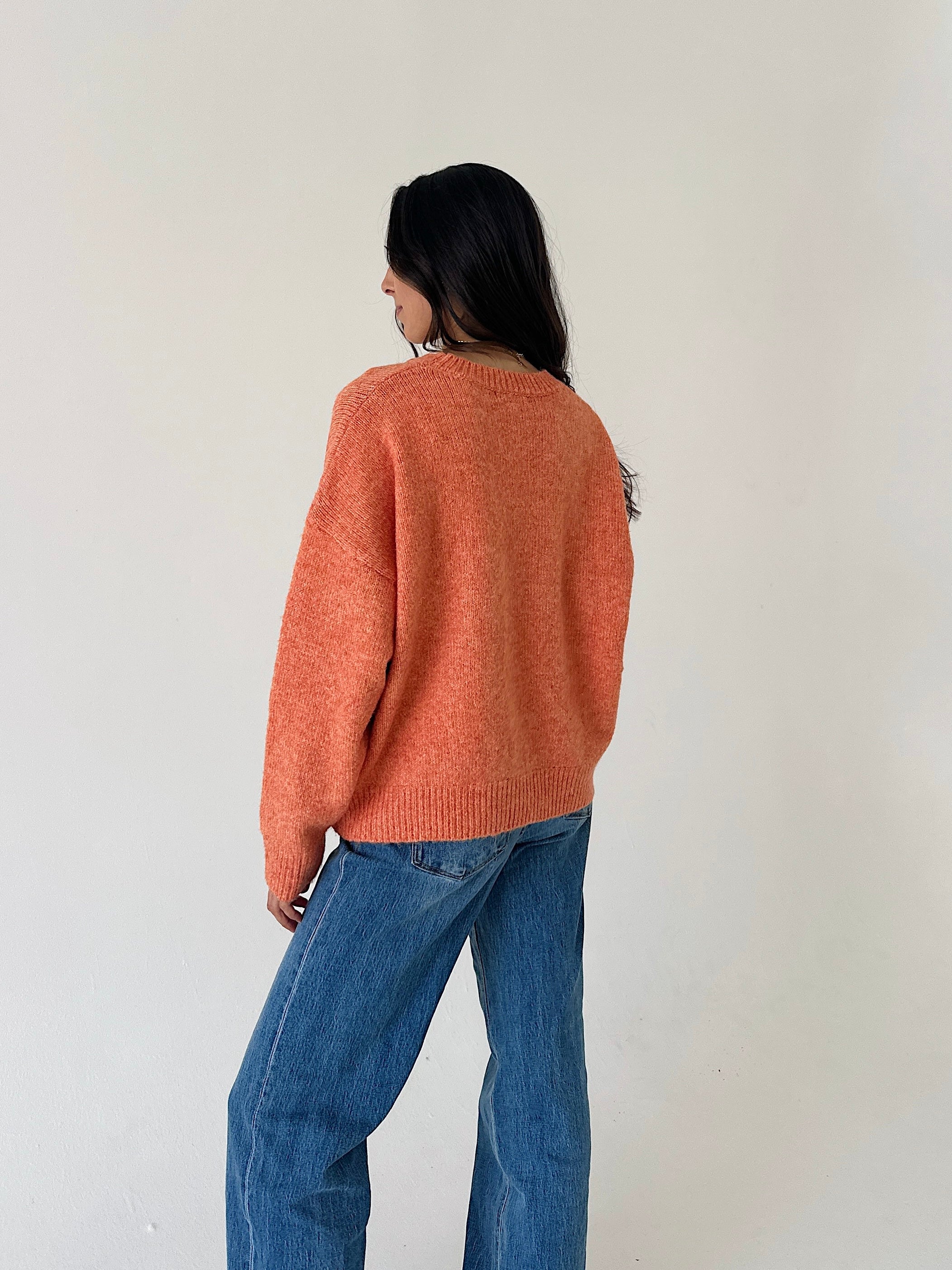 A woman wearing a fuzzy orange crewneck sweater and wide-leg blue jeans, photographed from behind against a plain off-white wall.
