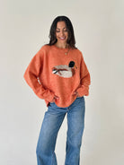 A woman wearing a fuzzy orange crewneck sweater with a knitted duck design on the front, paired with blue jeans, standing against a plain off-white wall.