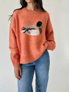 A woman wearing a fuzzy knit crewneck sweater in a peachy orange color, featuring a large duck motif across the chest, paired with blue jeans.