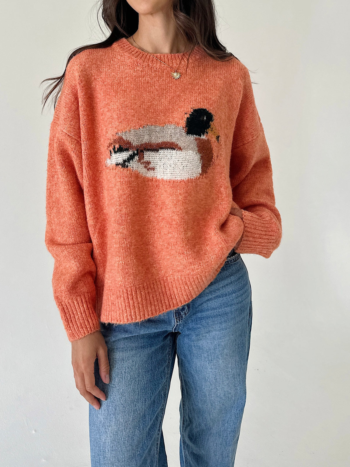 A woman wearing a fuzzy knit crewneck sweater in a peachy orange color, featuring a large duck motif across the chest, paired with blue jeans.