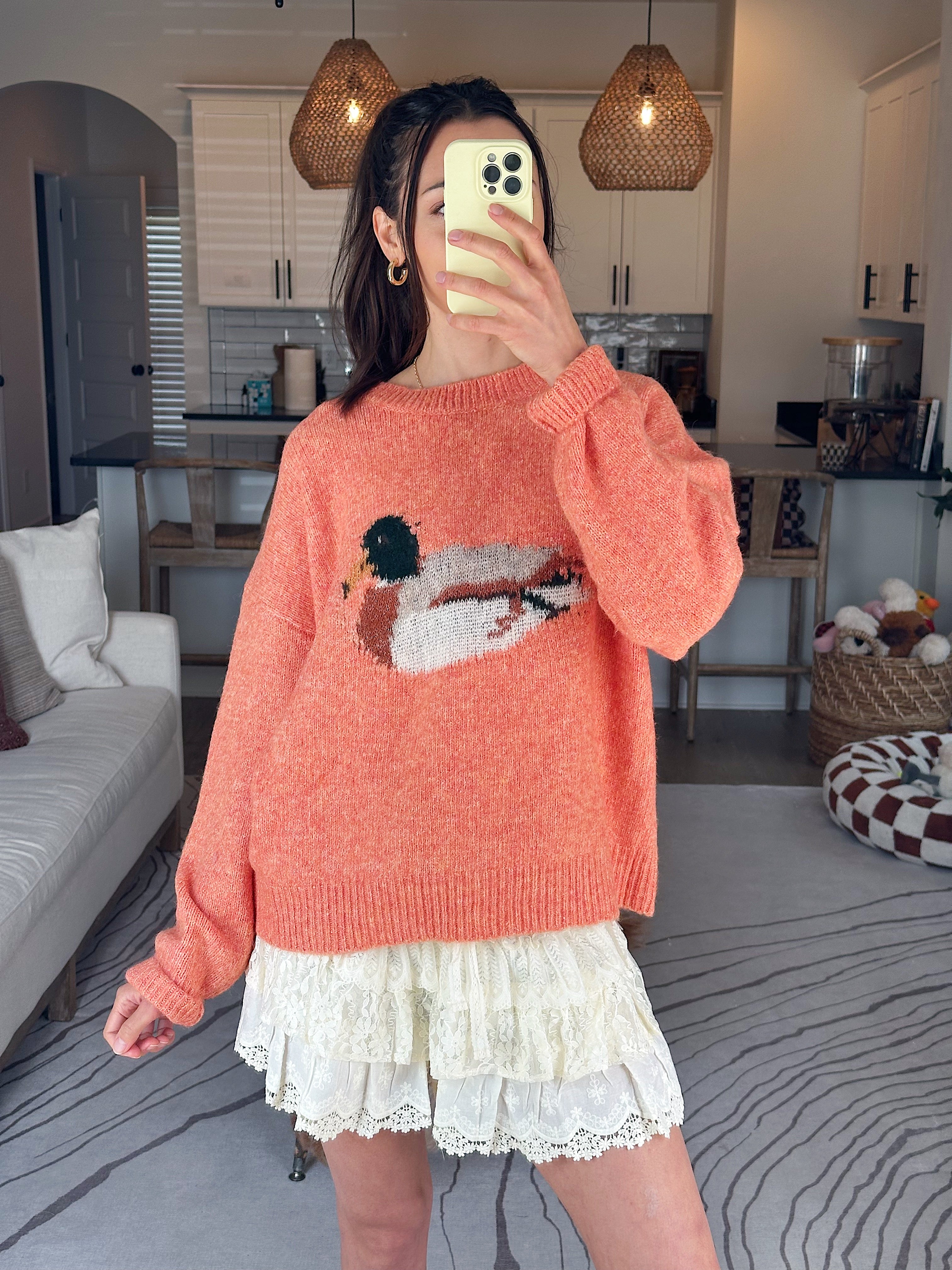 Person wearing an orange sweater with a graphic design, taking a mirror selfie in a room.