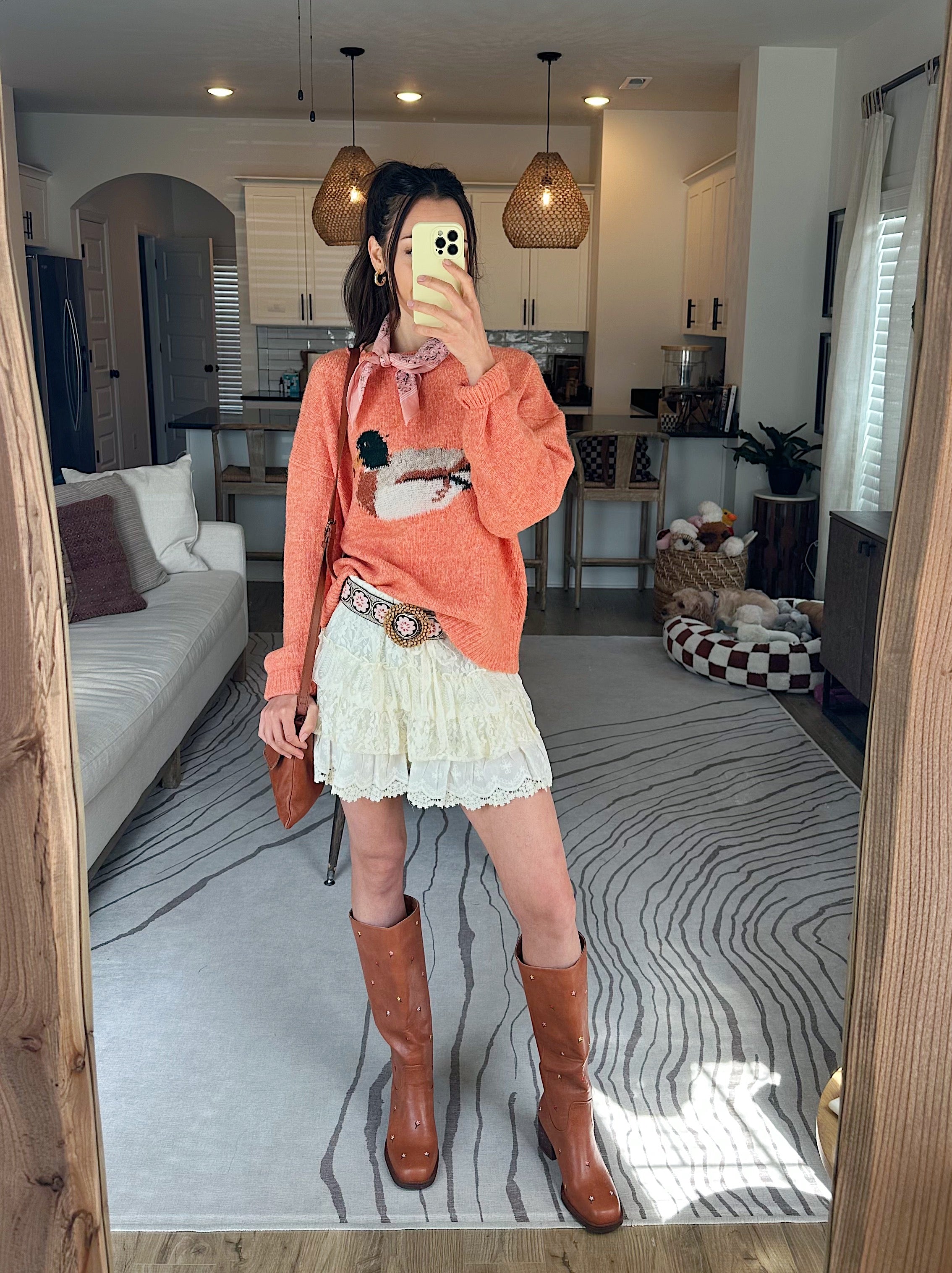Person taking a mirror selfie in a living room wearing an orange sweater, white skirt, and brown boots.