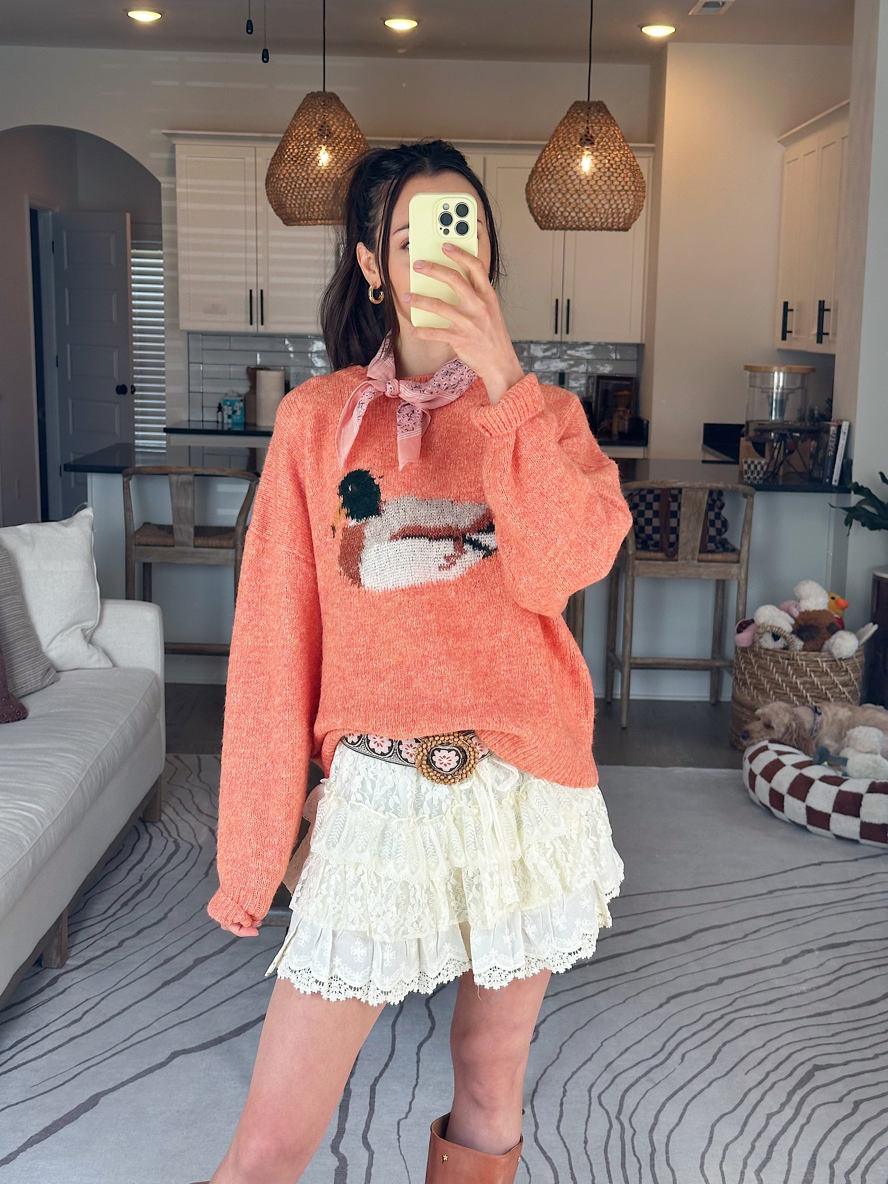Person wearing an orange sweater with a duck design and a white skirt in a living room.