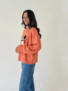 A woman wearing a fuzzy orange crewneck sweater with a small embroidered character on the chest, paired with blue jeans, standing against a plain off-white wall.
