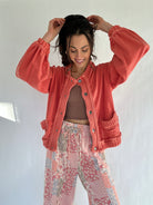 Woman wearing a coral jacket and floral pants against a white background putting her hair up