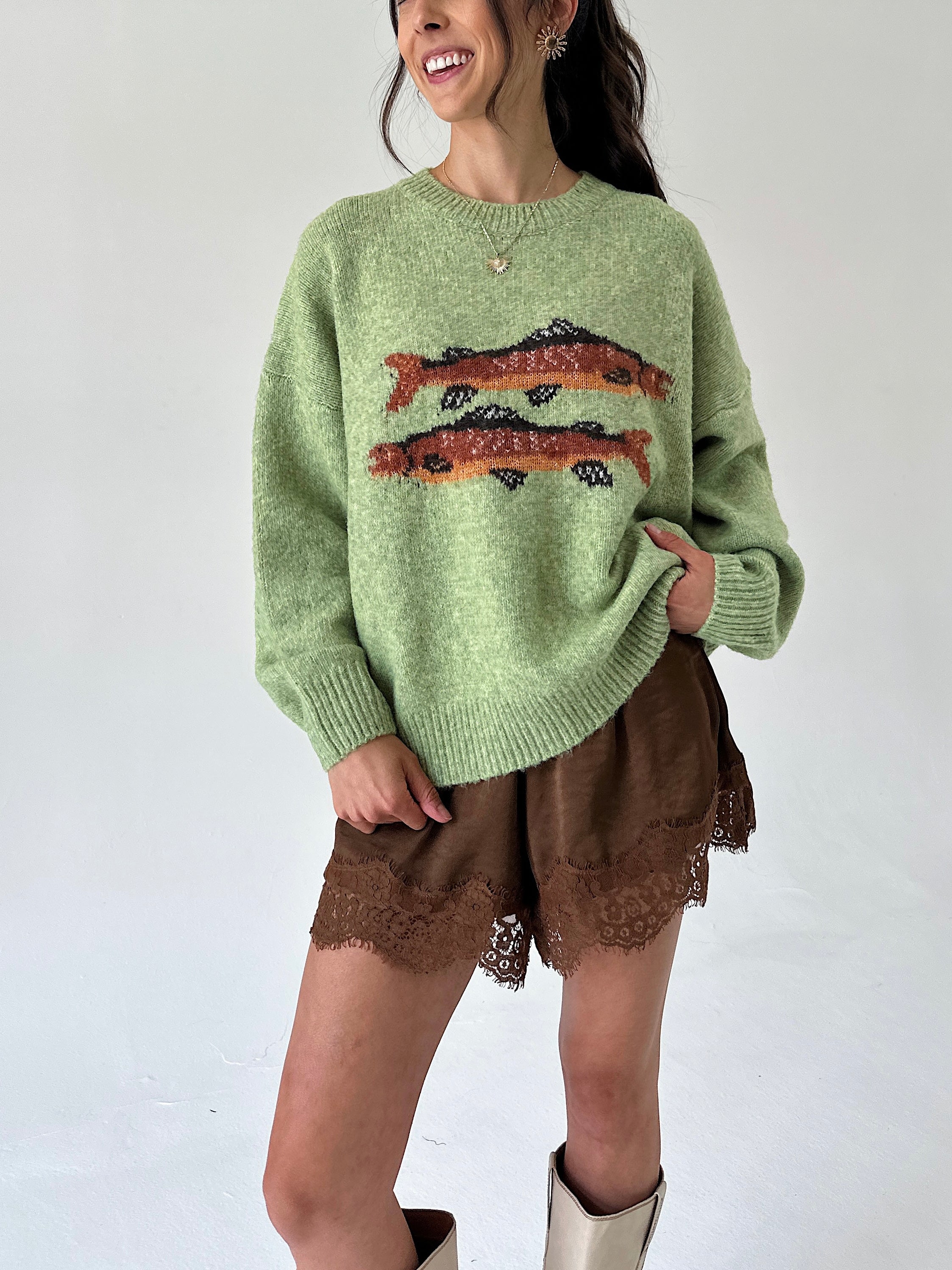 A woman wearing a light green knit sweater with two orange and black fish motifs across the chest, paired with brown lace-trim shorts and beige boots, against a plain white background.