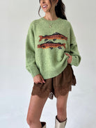 A woman wearing a light green knit sweater with two orange and black fish motifs across the chest, paired with brown lace-trim shorts and beige boots, against a plain white background.