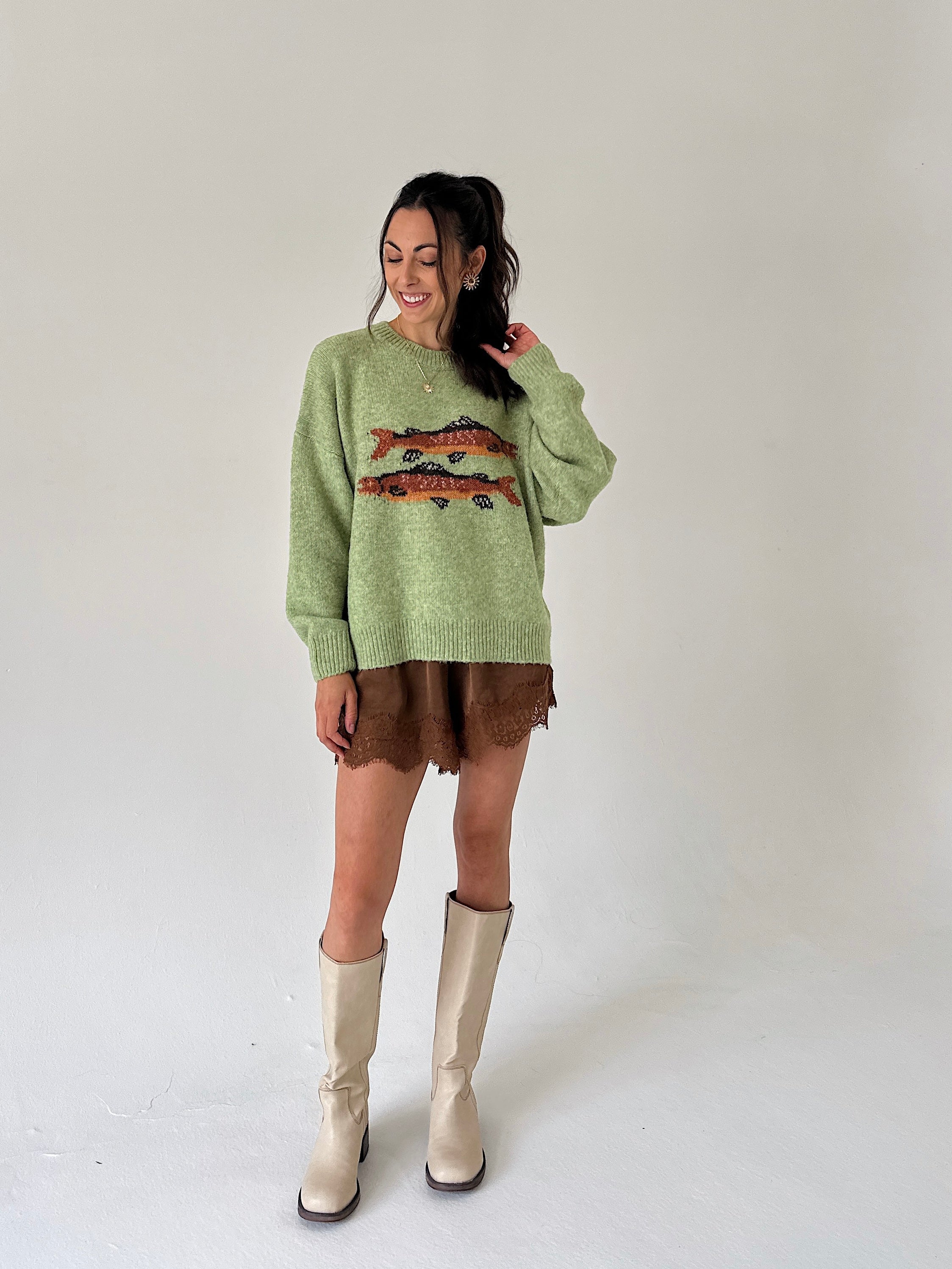 A woman modeling a light sage green knit sweater with a colorful fish graphic across the chest, paired with a brown satin shorts and beige knee-high boots, against a plain light gray studio background.