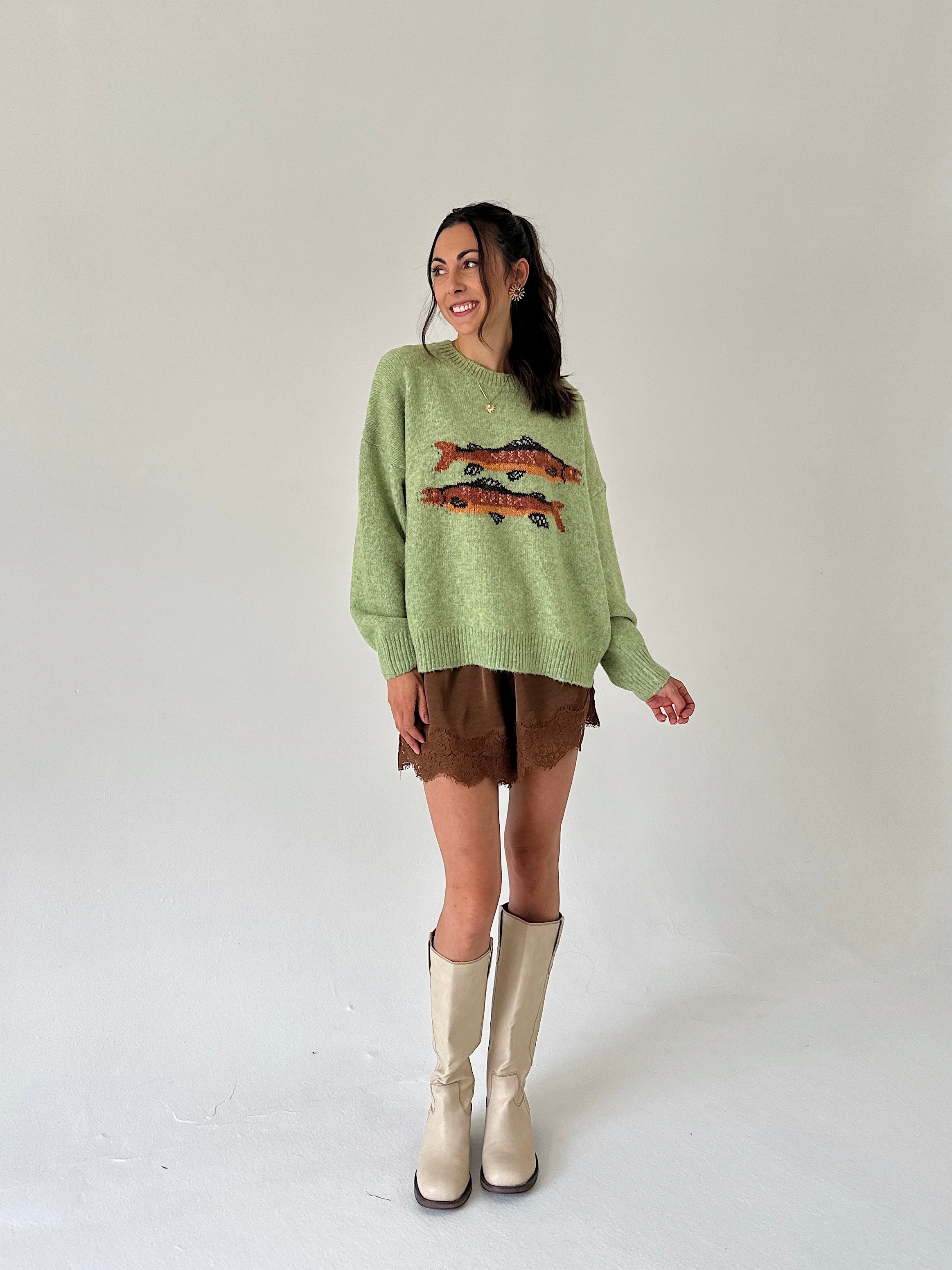 A woman modeling a light green knit sweater with a fish graphic, paired with brown shorts and beige knee-high boots, against a plain light gray studio background.