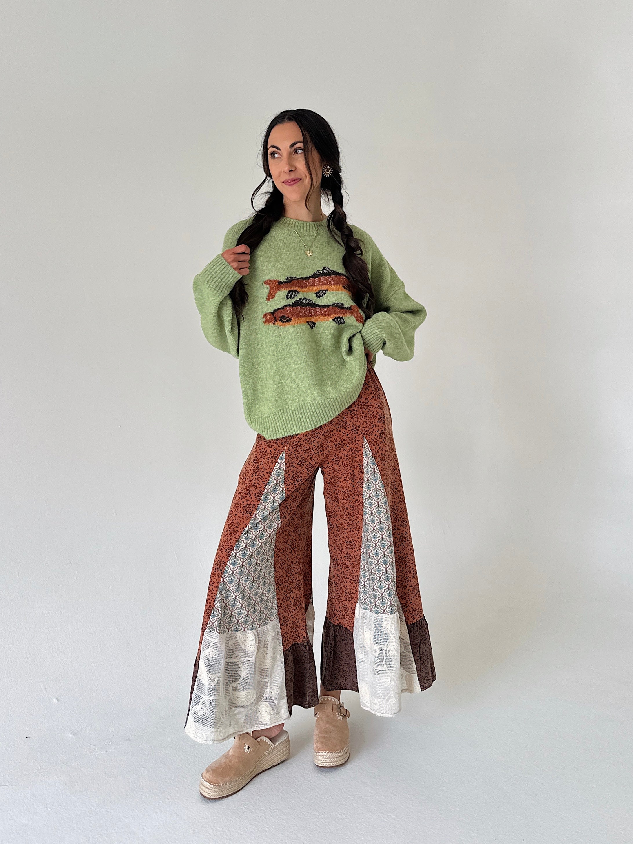 A woman modeling a light green fuzzy knit sweater with a colorful trout graphic, paired with rust-brown wide-leg patterned pants featuring lace accents and beige slip-on shoes, against a plain light gray studio background.