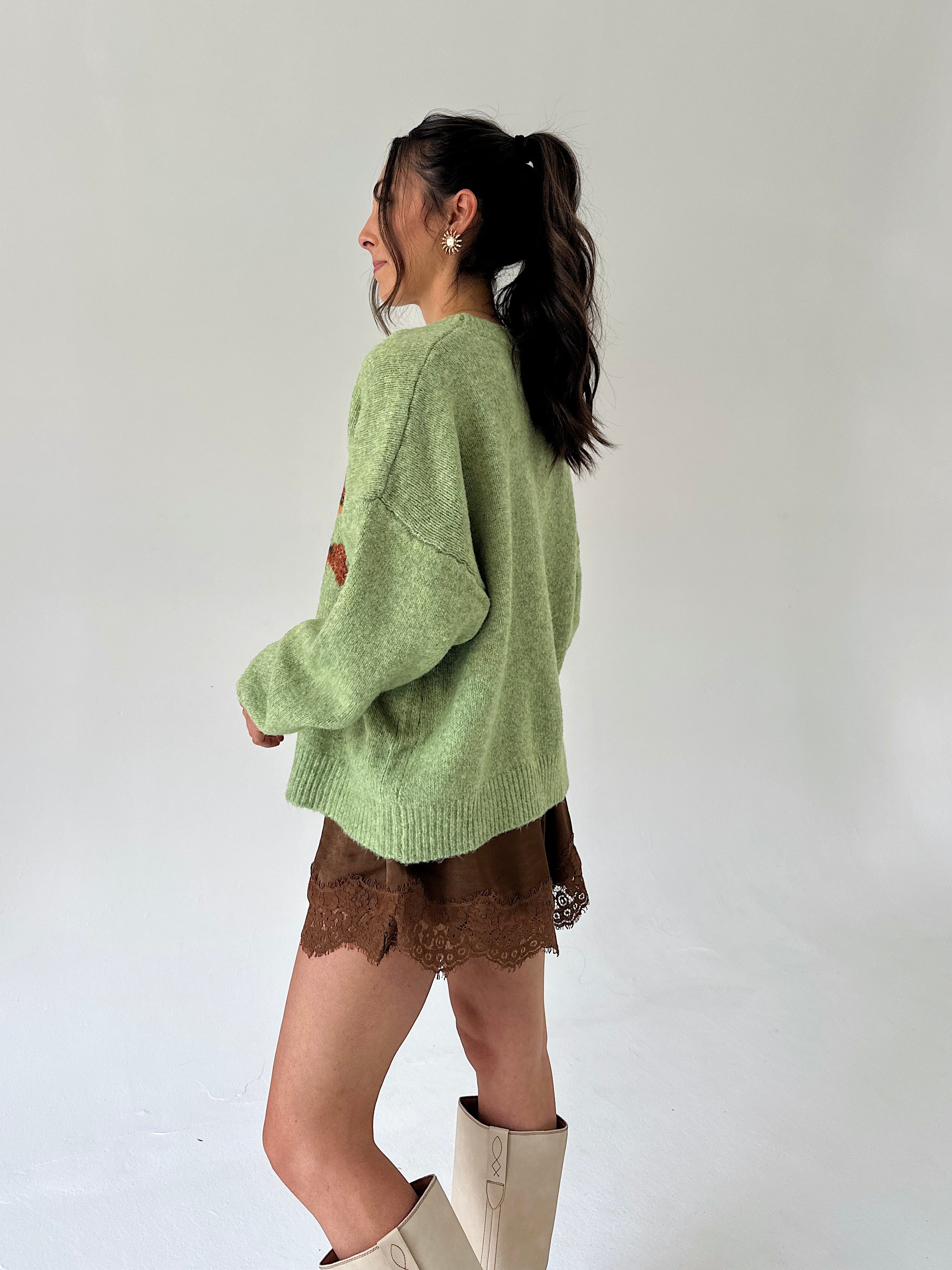 A woman wearing a light sage green knit sweater, brown lace shorts, and beige boots, photographed from the side against a plain white studio background.