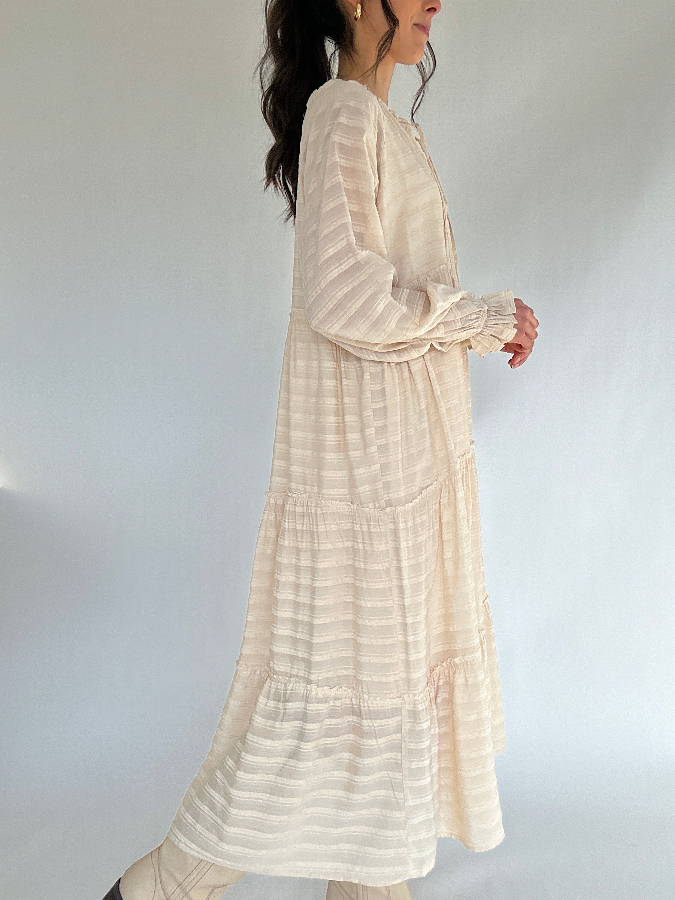 Woman wearing a long, beige-colored dress against a plain background from side