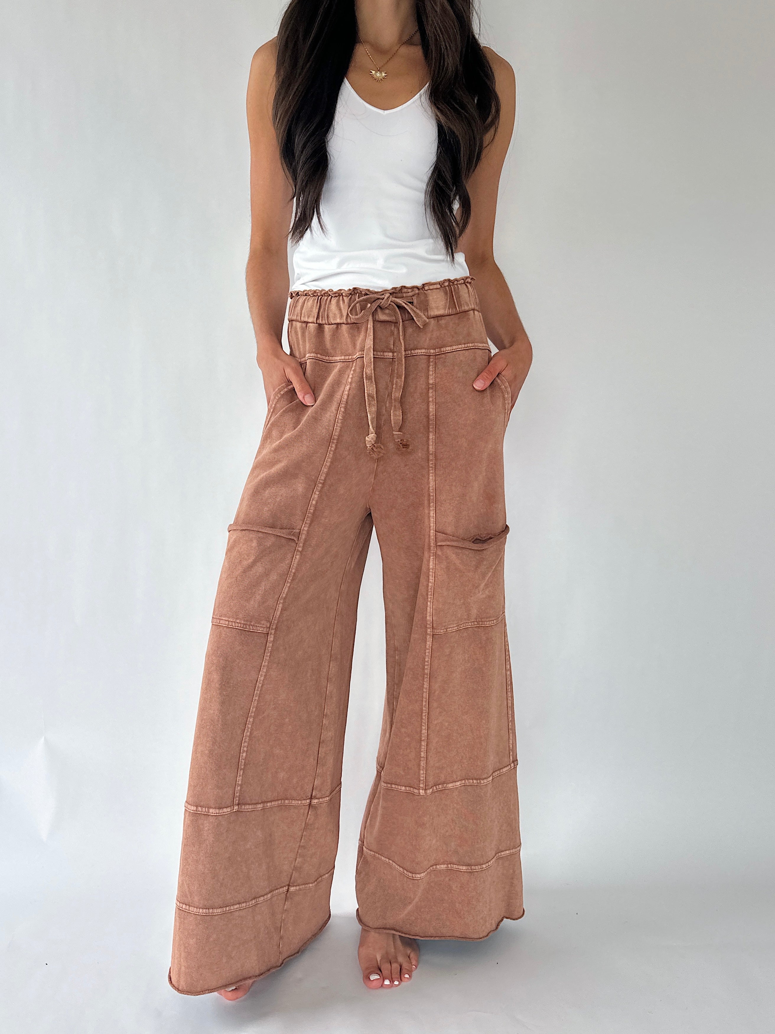 A woman wearing wide-leg, high-waisted pants in a dusty rose-brown  with a drawstring waist and front patch pockets, paired with a white tank top, standing against a light gray studio background.