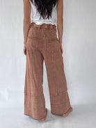 A person shown from the back wearing high-waisted, wide-leg brown pants with a ruffled waistband, a white sleeveless top, and bare feet against a plain light background.