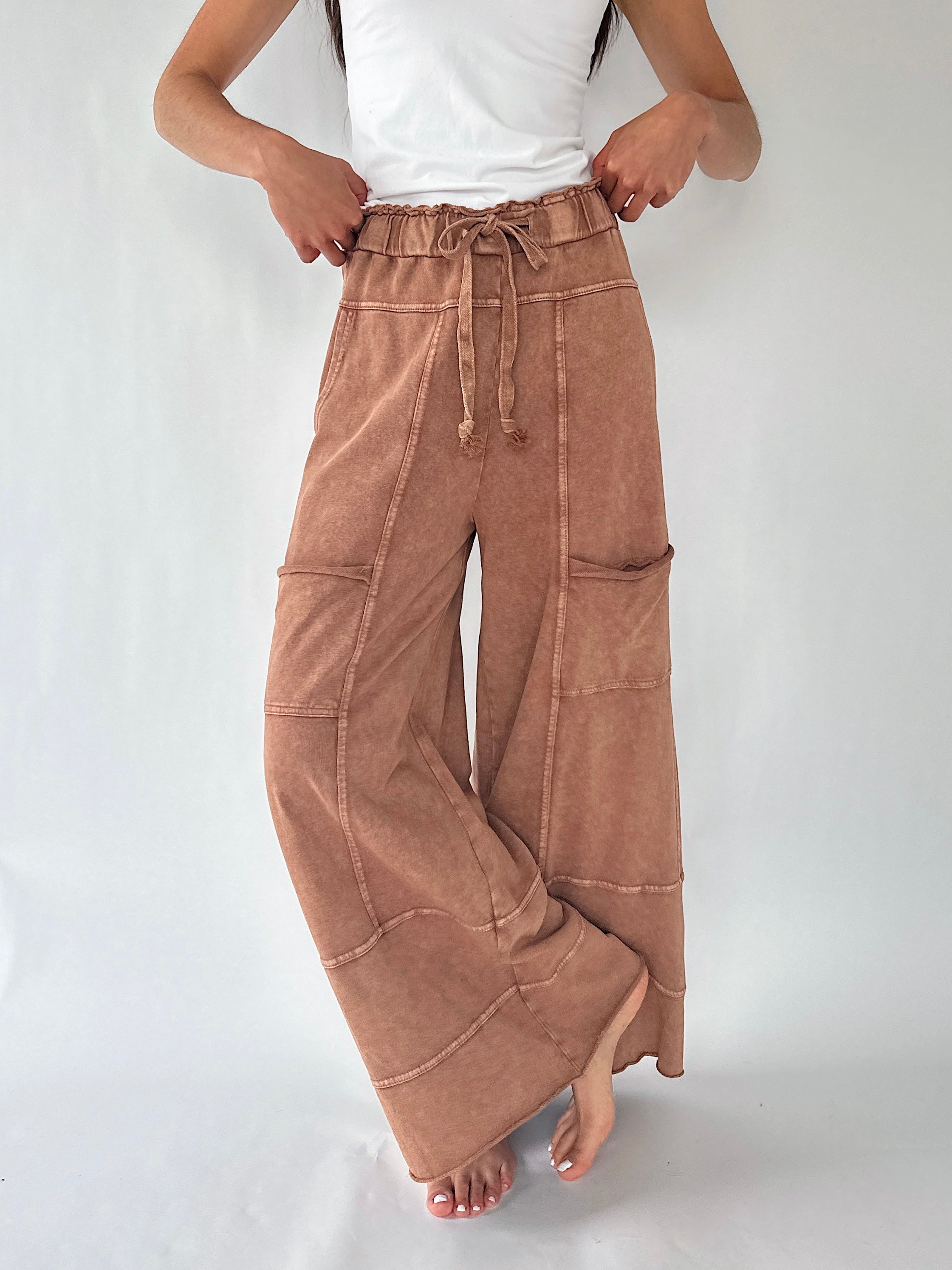 A person wearing wide-leg brown pants with a drawstring waistband and large patch pockets, paired with a white sleeveless top, standing against a plain light background.